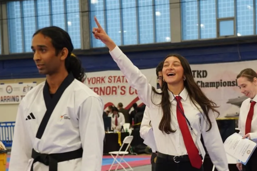 Teen girl smiling and pointing upward during taekwondo competition, with other competitors in background