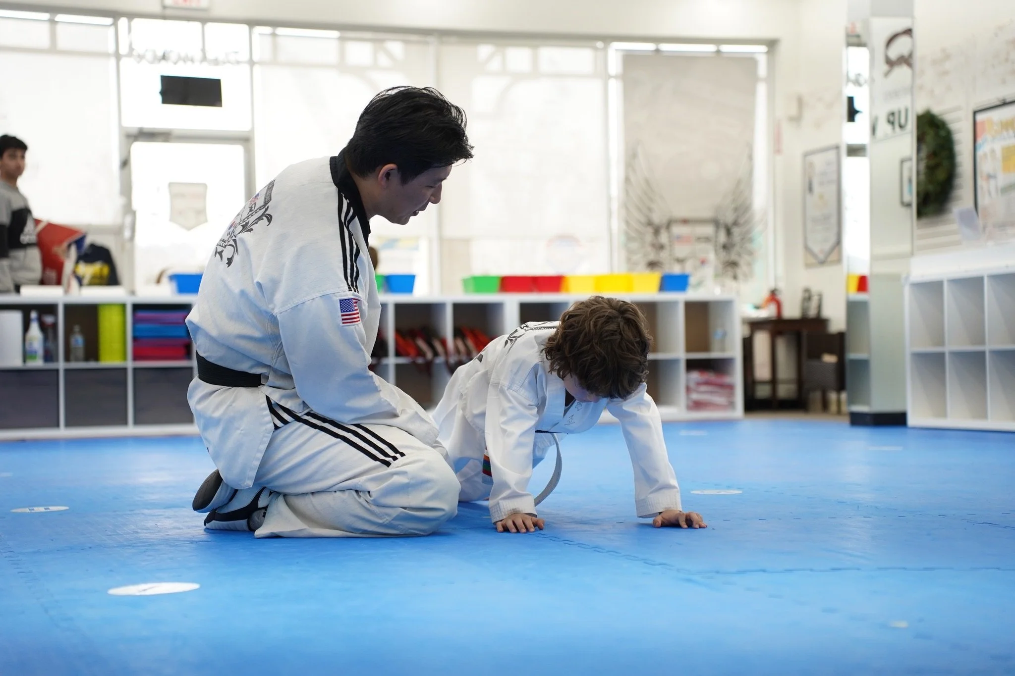 An adult and a child practicing at a martial arts dojo, with the adult kneeling and helping the child on the blue mat.