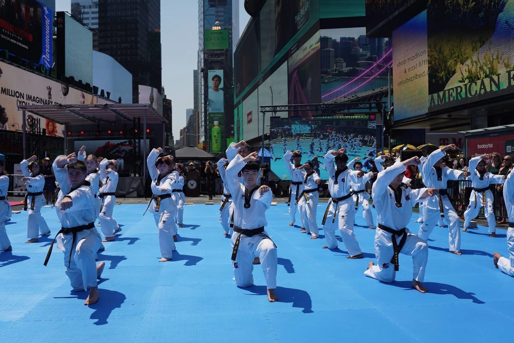 Group of martial artists practicing taekwondo outdoors on a blue mat in a city square with large electronic billboards and skyscrapers in the background.