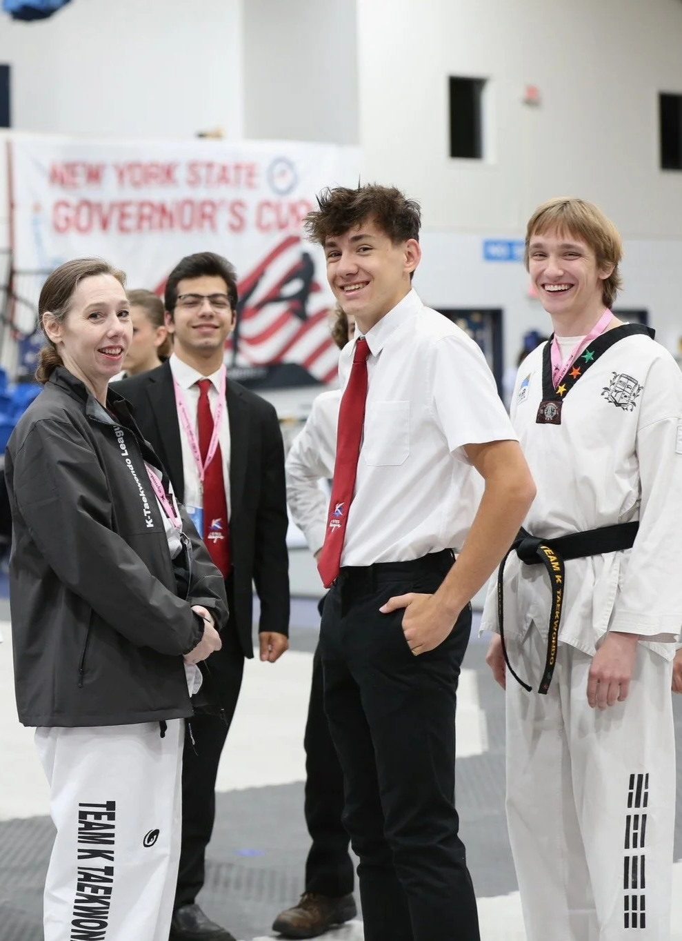 Group of five young people smiling at a martial arts event in a gymnasium, with a banner reading 'New York State Governor's Cup' in the background.