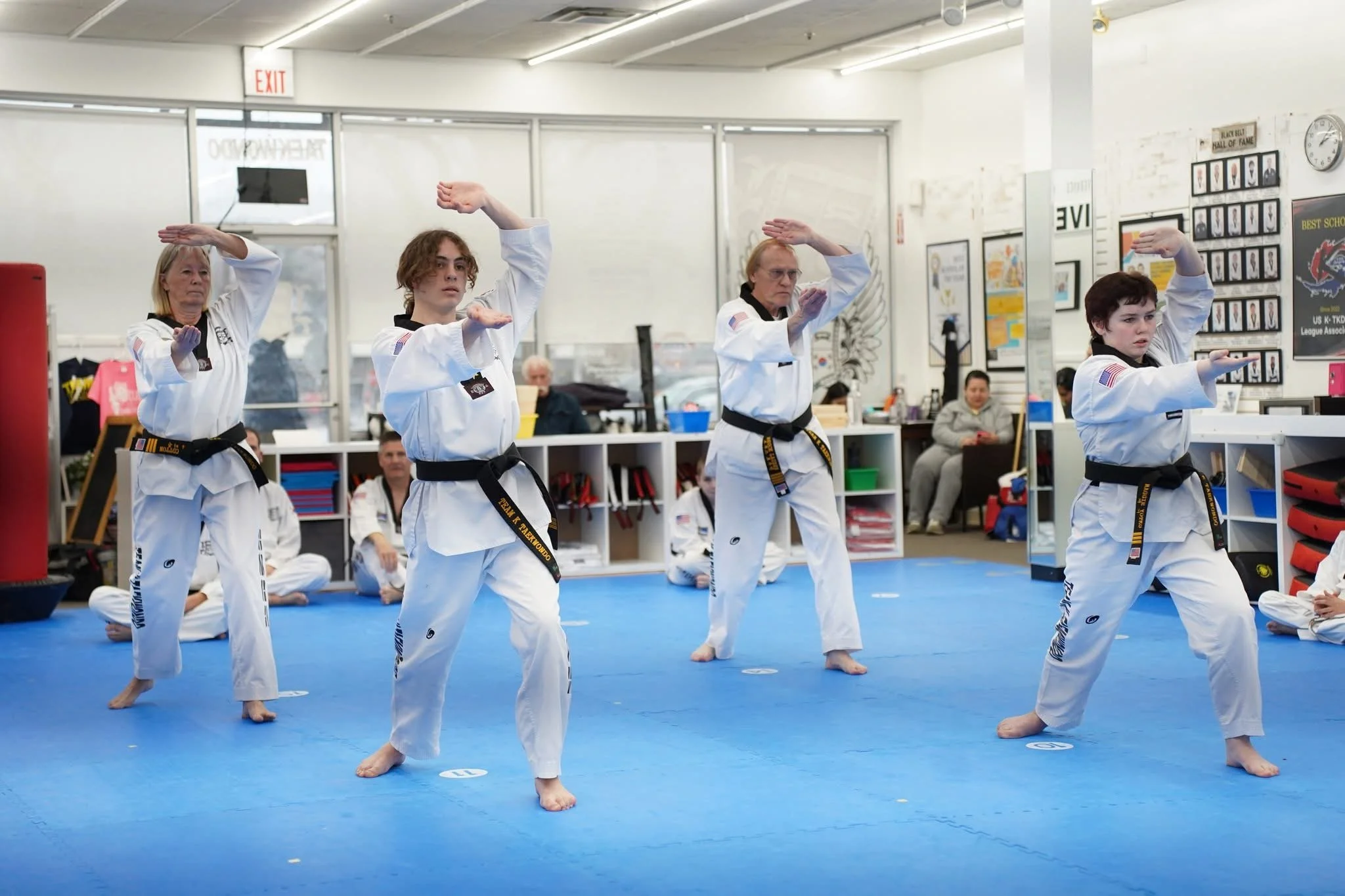 Martial arts class with students practicing moves in a gym, wearing white uniforms with black belts, on a blue mat with student numbers.