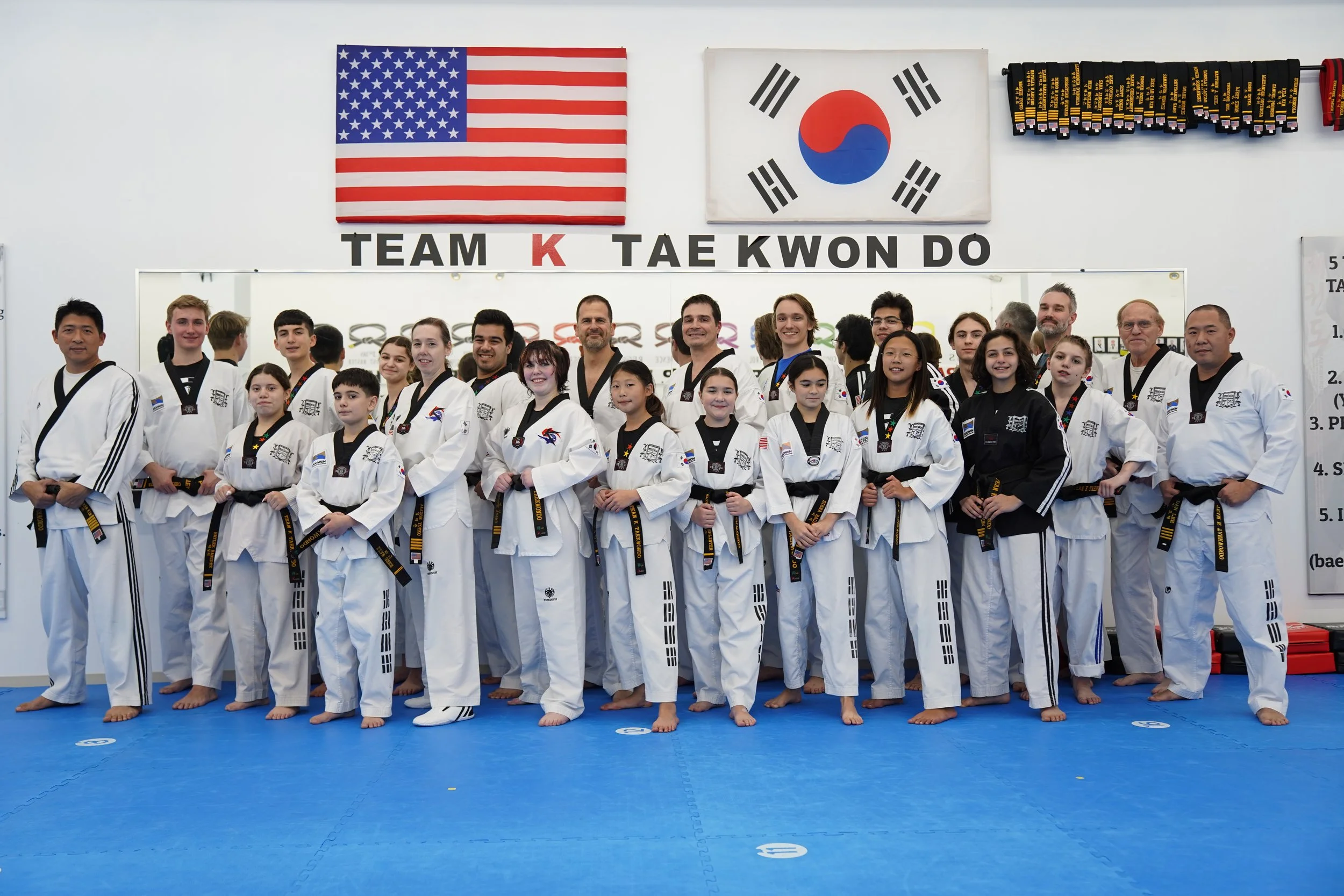 Group of martial arts students and instructors in uniform posing in a martial arts dojo, with flags of the United States and South Korea on the wall behind them.