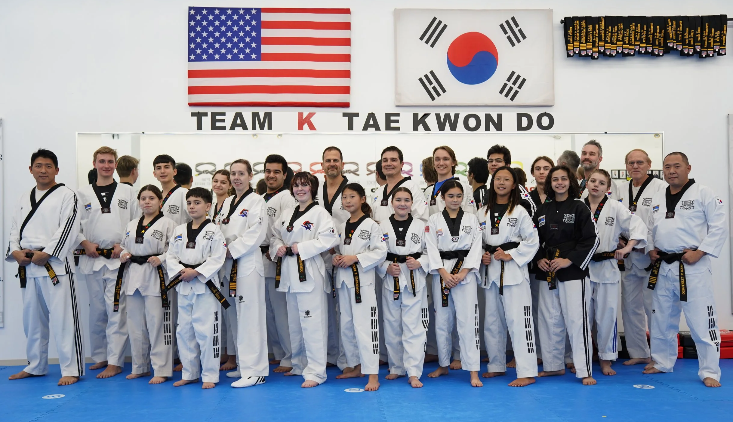 Group of taekwondo practitioners in white and black uniforms posing for a photo in a taekwondo training facility with American and South Korean flags.