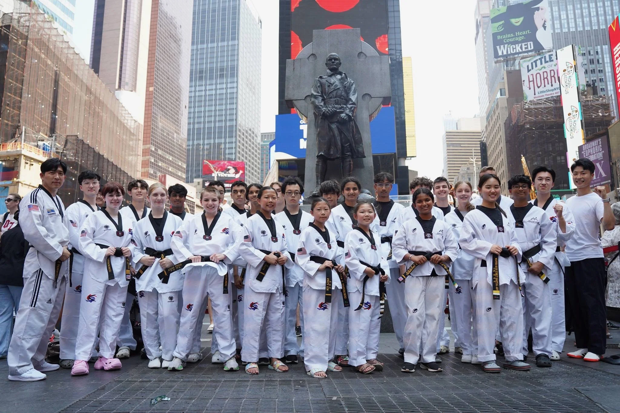 Group of children and young adults in taekwondo uniforms standing in Times Square, New York City, with billboards and tall buildings in the background.