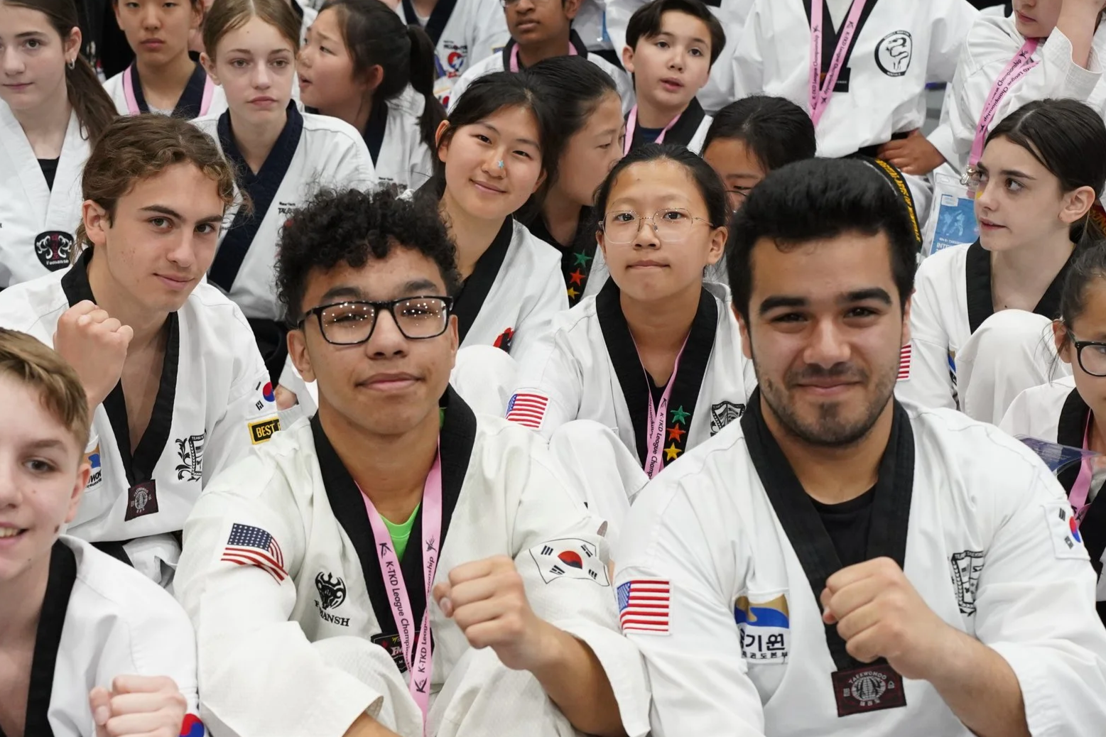Group of children and teenagers in martial arts uniforms at a taekwondo event, some making fist gestures, wearing badges and medals.