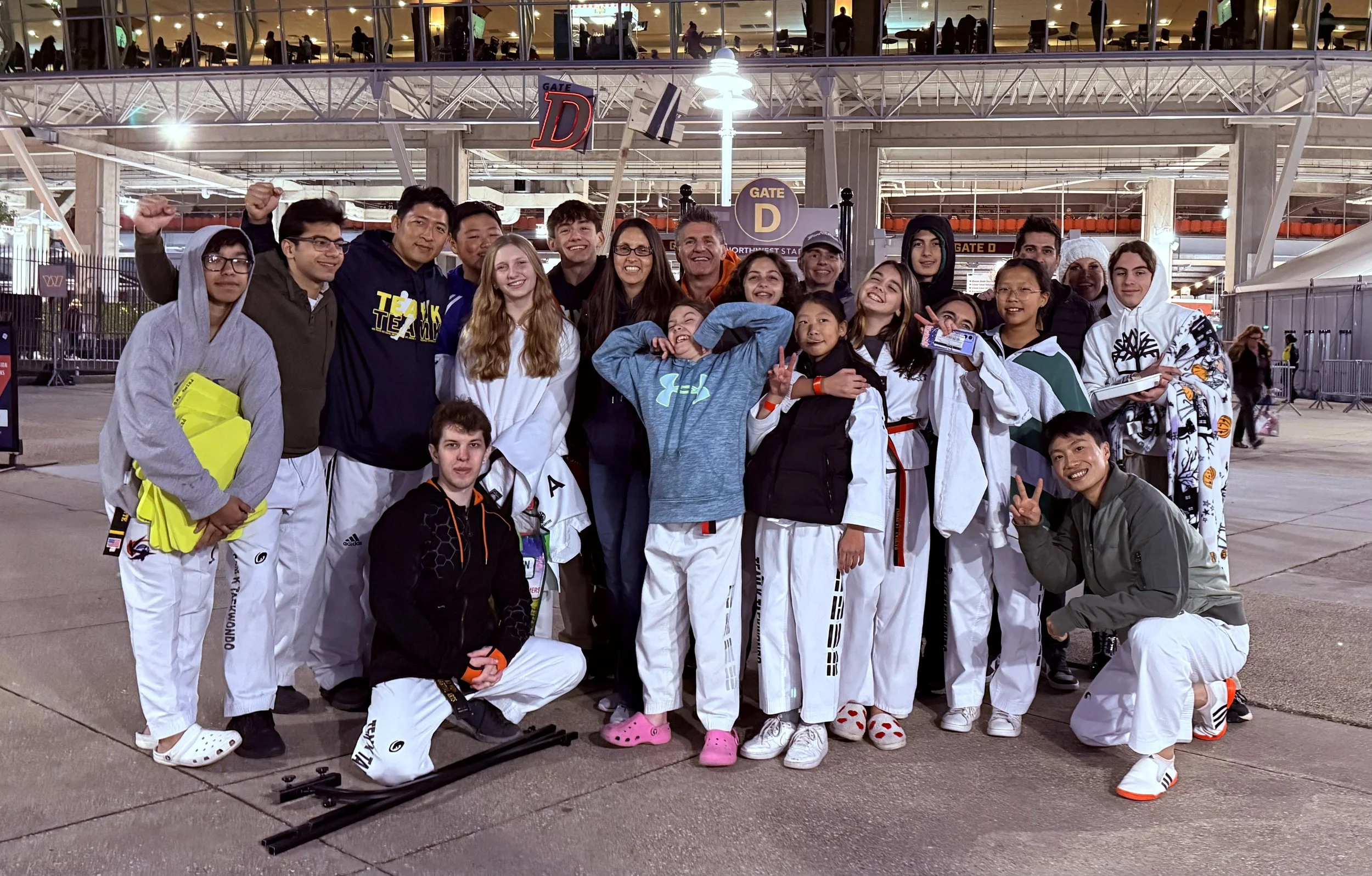 Group of athletes in martial arts uniforms posing at an airport, some smiling, making peace signs, some raising fists, with luggage and airport signs visible in the background.