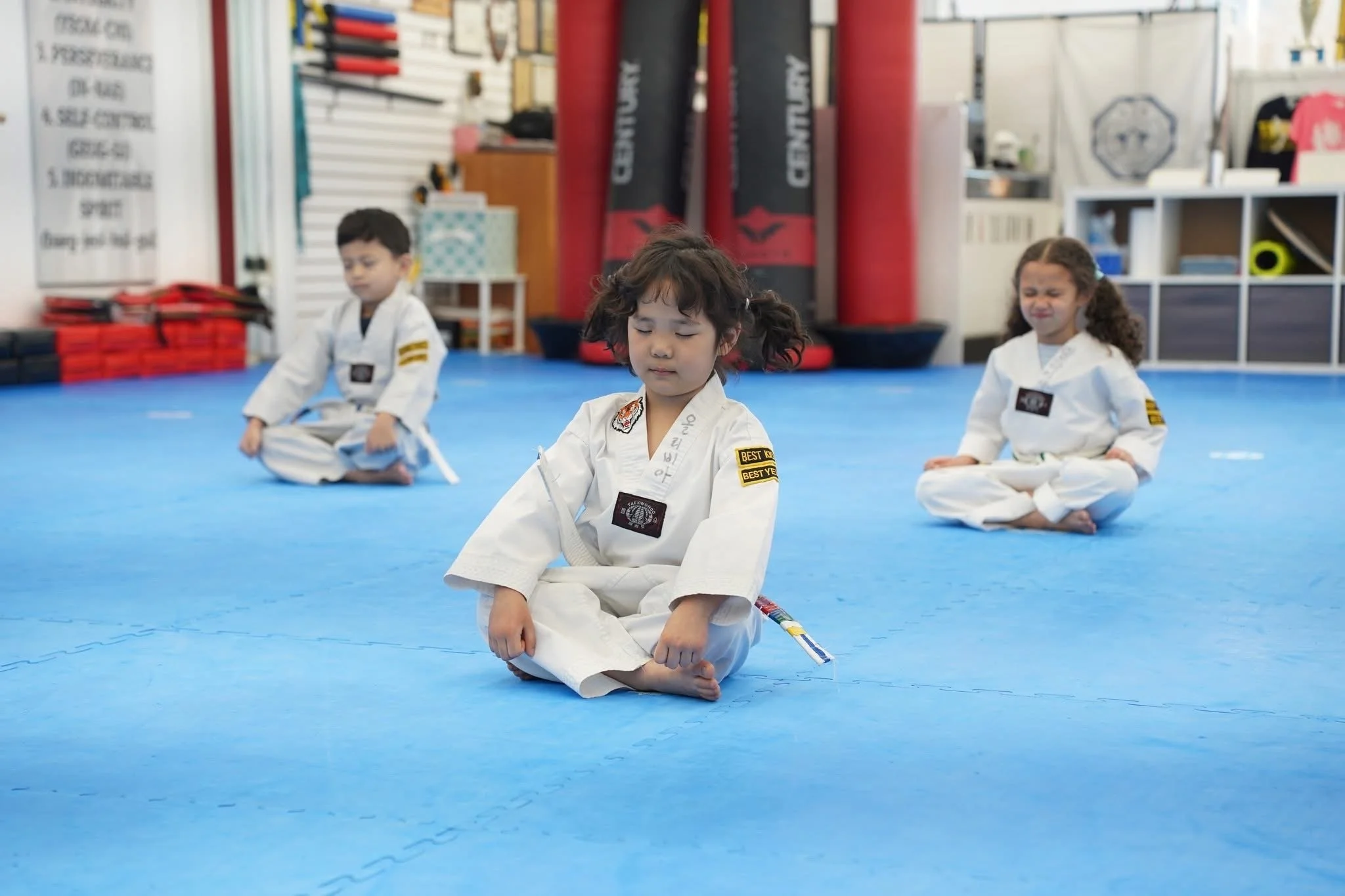 Three children dressed in martial arts uniforms meditating in a dojo with a blue padded floor.
