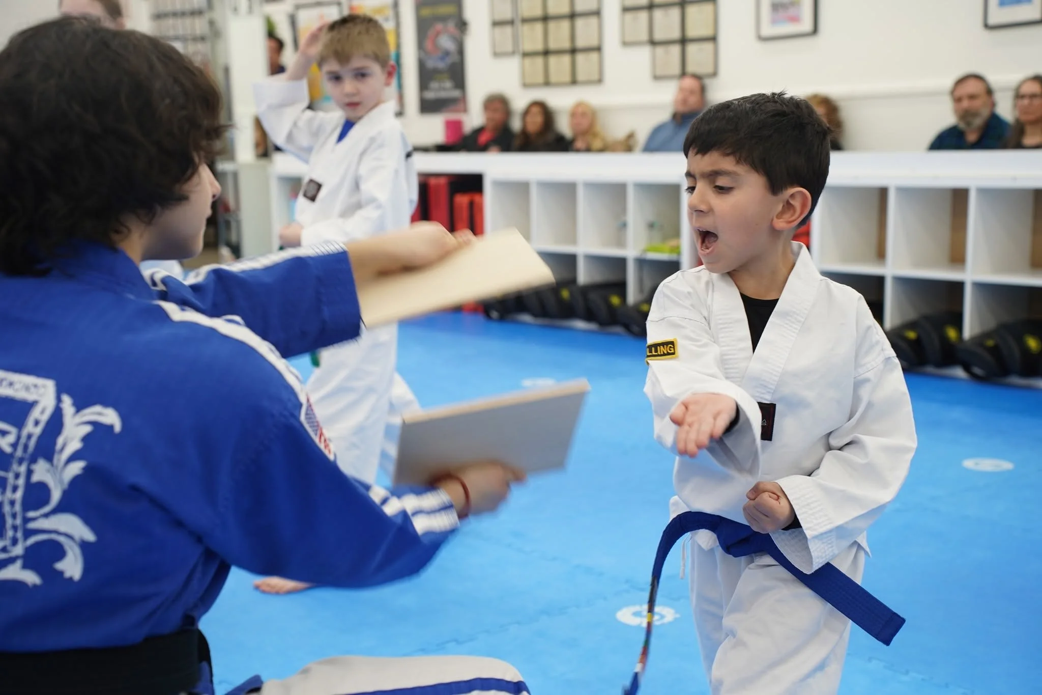 Two young boys in martial arts uniforms exchanging a handshake or bow in a martial arts studio, with a group of spectators seated in the background.