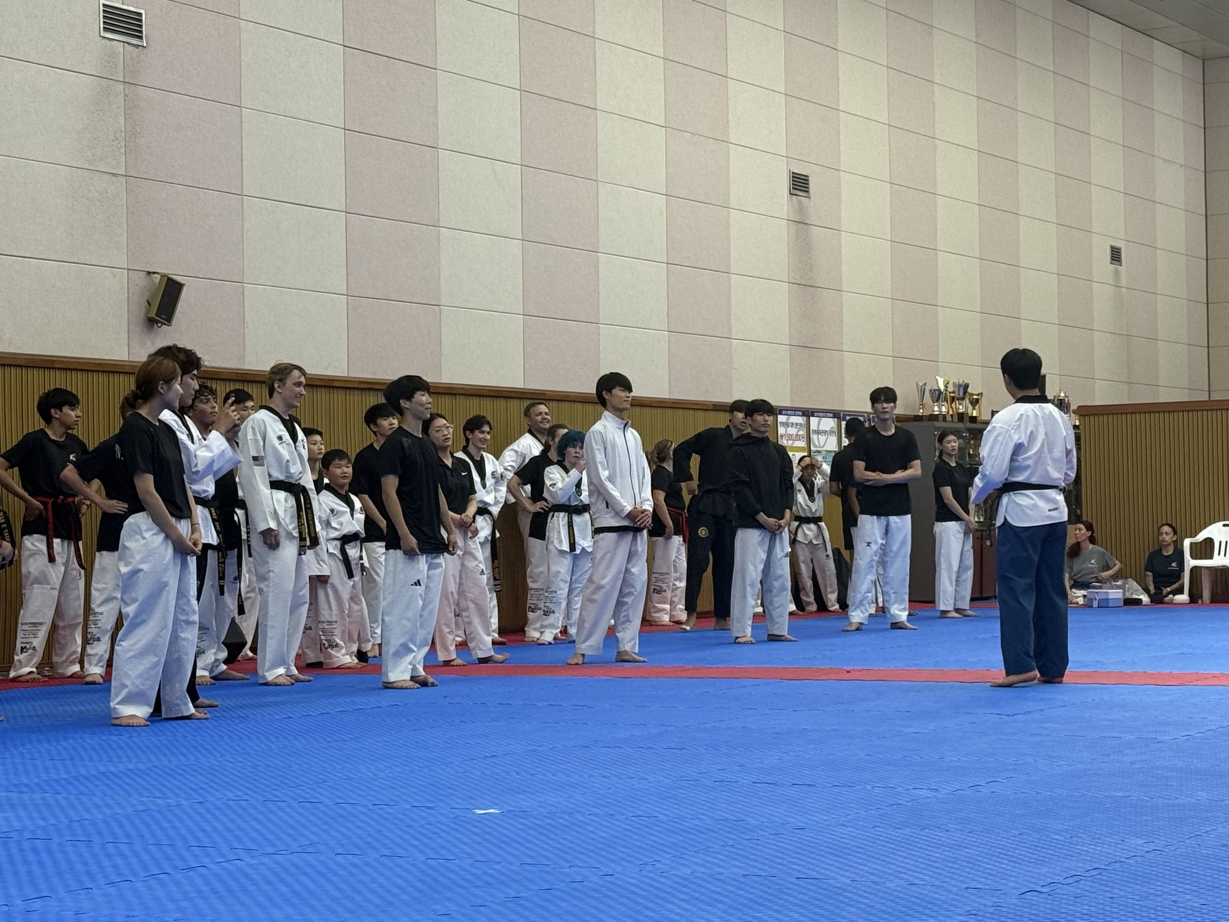 Martial arts students dressed in traditional uniforms, standing in a line on a blue mat, listening to an instructor in a white uniform during a competition or class in a gymnasium