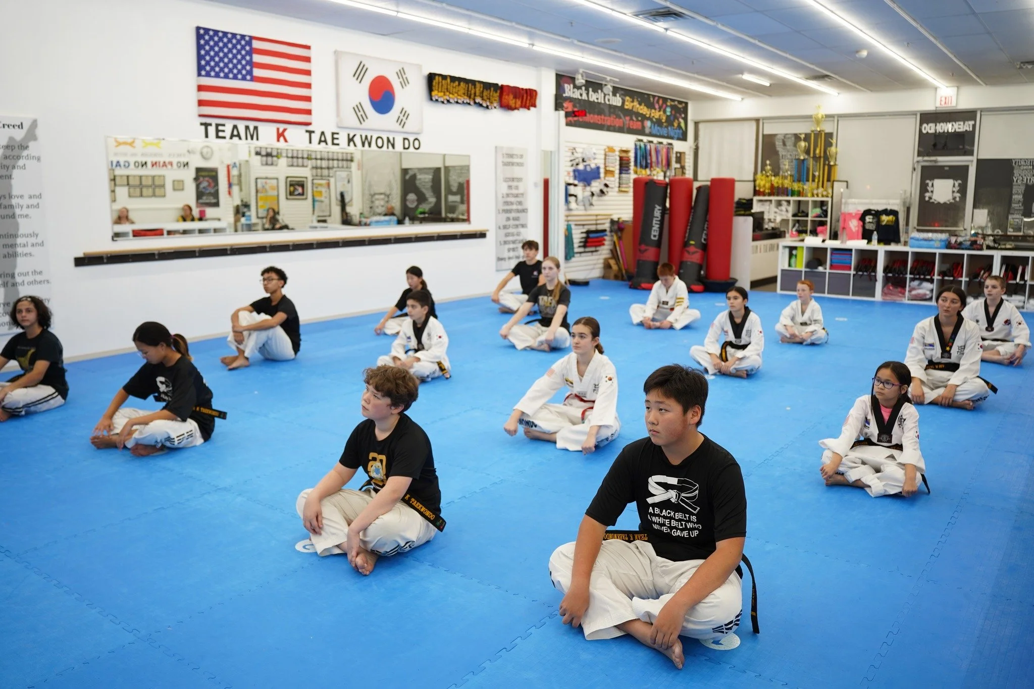 Children and teenagers seated on the blue mat floor in a martial arts gym, with flags of the United States and South Korea and martial arts banners on the wall behind them.