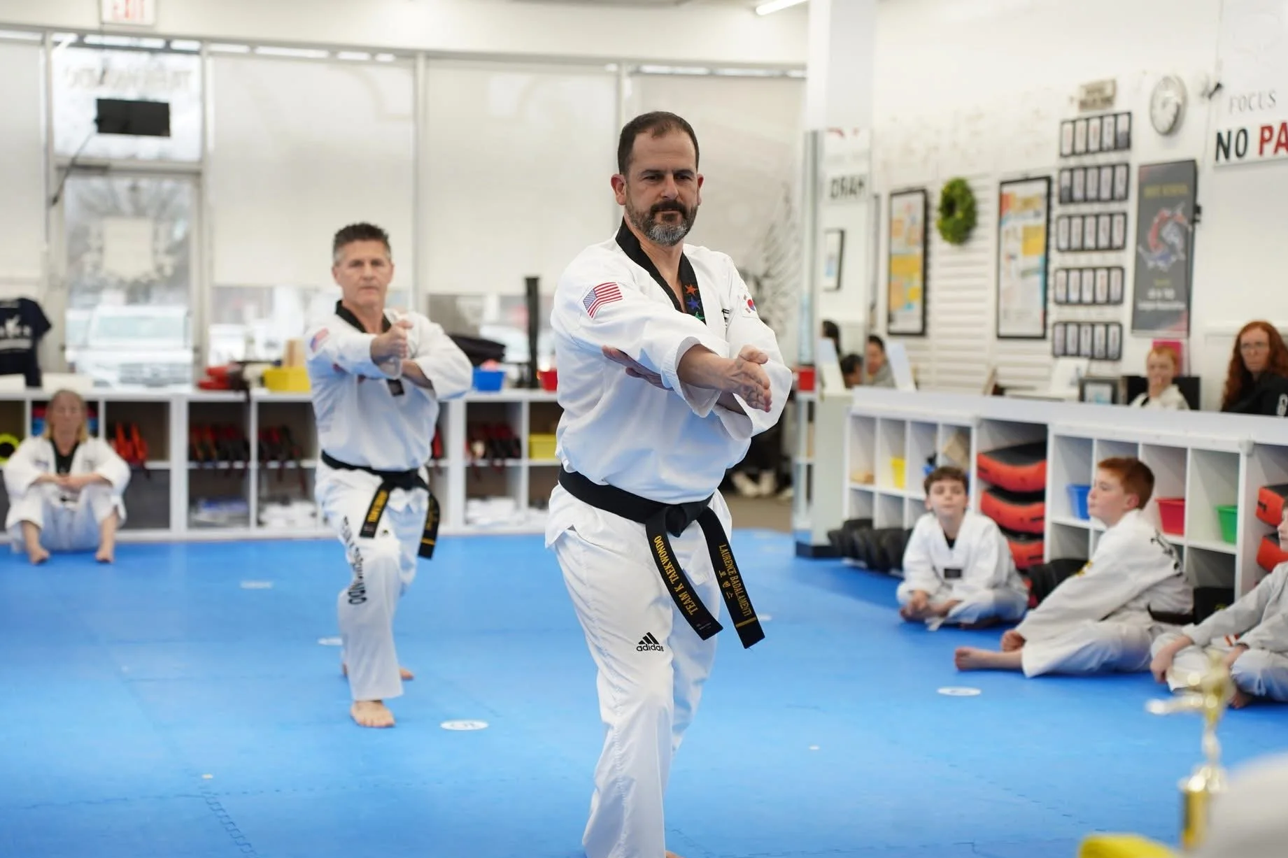Taekwondo class with instructor performing a kick in the foreground, students sitting and watching in a martial arts dojo.