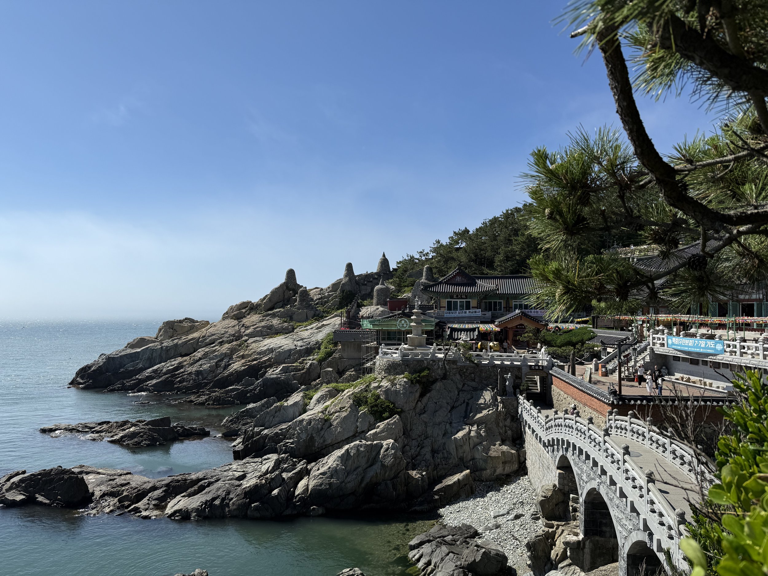 A view of a seaside temple complex with traditional architecture, surrounded by trees, rocky shoreline, and overlooking clear water under a bright blue sky.