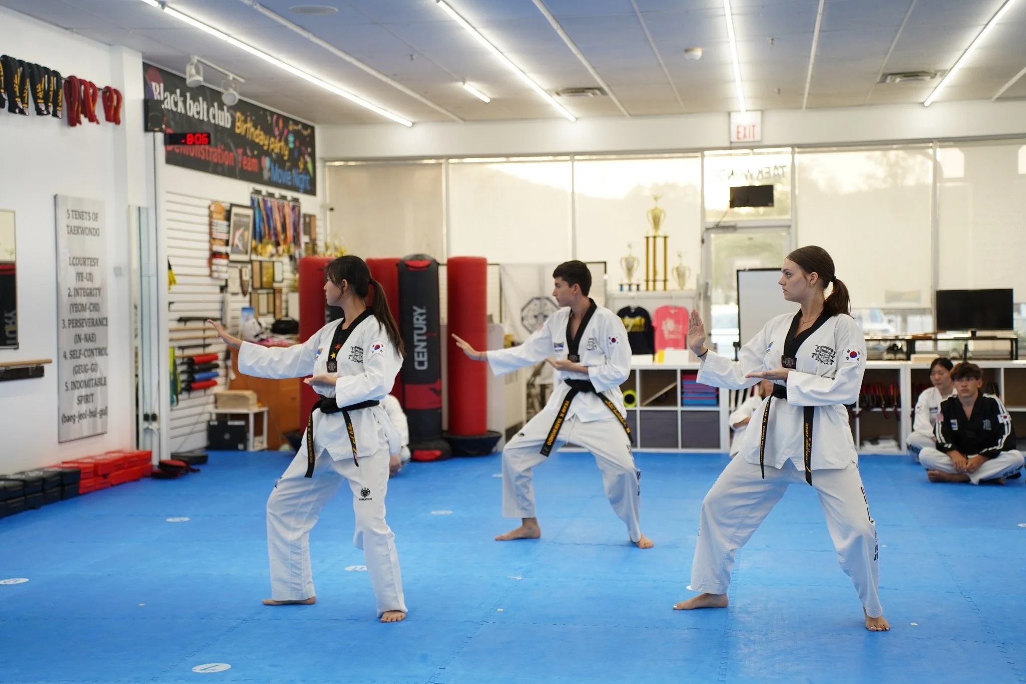 Three martial arts students practicing Taekwondo in a dojo, wearing white uniforms with black belts, and one student in a black uniform sitting on the blue mat. There are martial arts equipment and trophies in the background.
