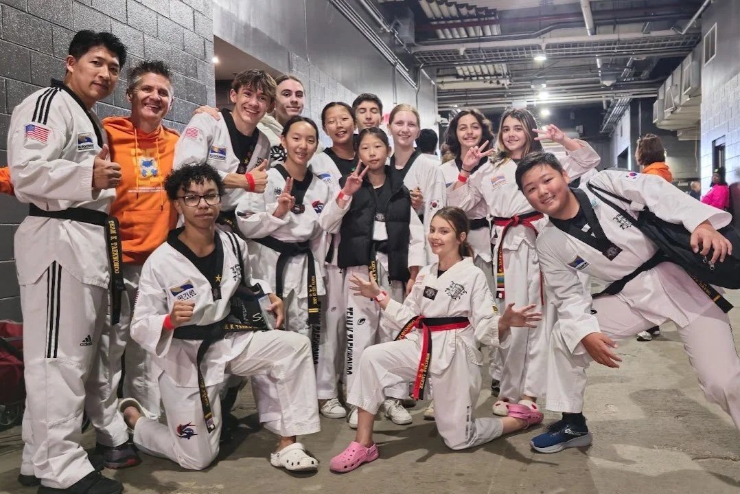 Group of young martial artists in white uniforms with various colored belts, posing with their instructor in an indoor facility, some making peace signs and smiling.
