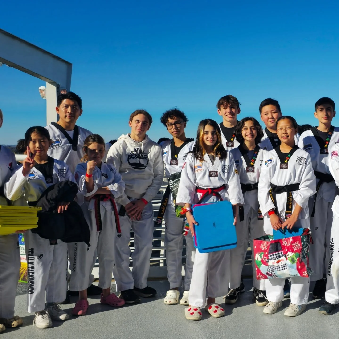 Group of children and teenagers in martial arts uniforms standing on a platform outdoors, smiling for the photo against a bright blue sky.