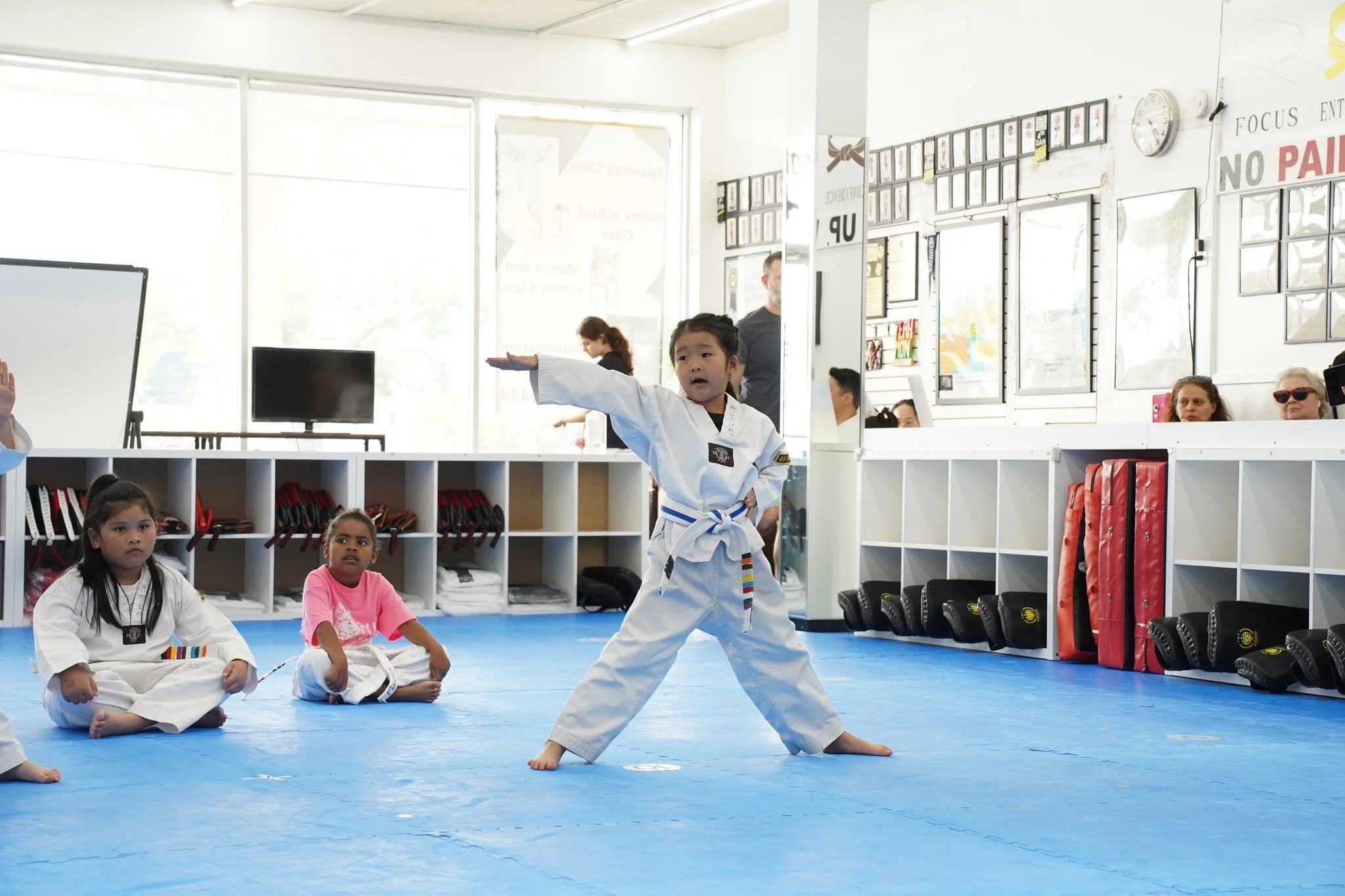 Young girl in a martial arts uniform practicing a stance in a martial arts class, with children sitting on the mat around her and adults watching in the background.