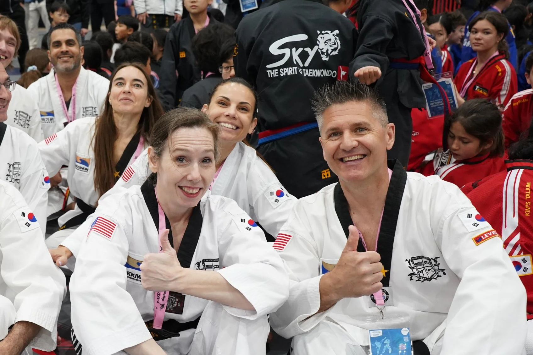 Group of taekwondo practitioners, including women and men in white uniforms with flags and patches, smiling and giving thumbs-up at a martial arts event surrounded by children in red uniforms.