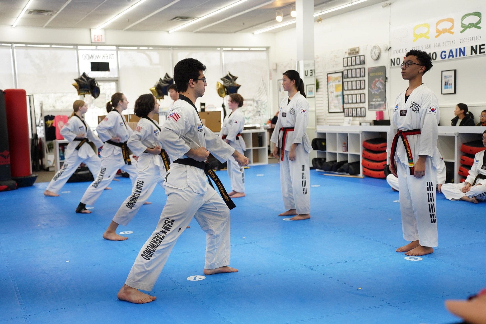 Martial arts class practicing traditional taekwondo techniques in a gym, with students wearing white uniforms and black belts, standing on a blue mat, facing an instructor.