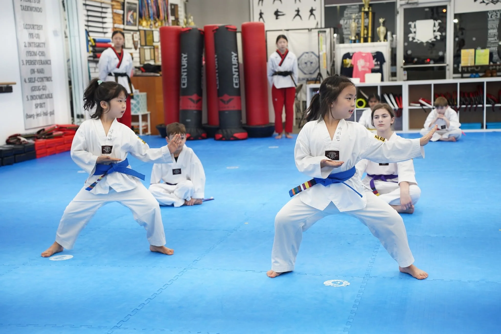 Young children practicing martial arts in a dojo, wearing white uniforms and colored belts, with an instructor and martial arts training equipment in the background.