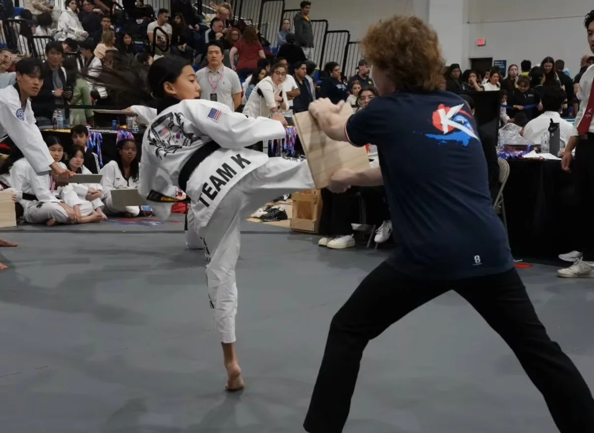 A young martial artist delivering a high kick to a wooden board held by an instructor in a martial arts competition, with an audience watching in the background.