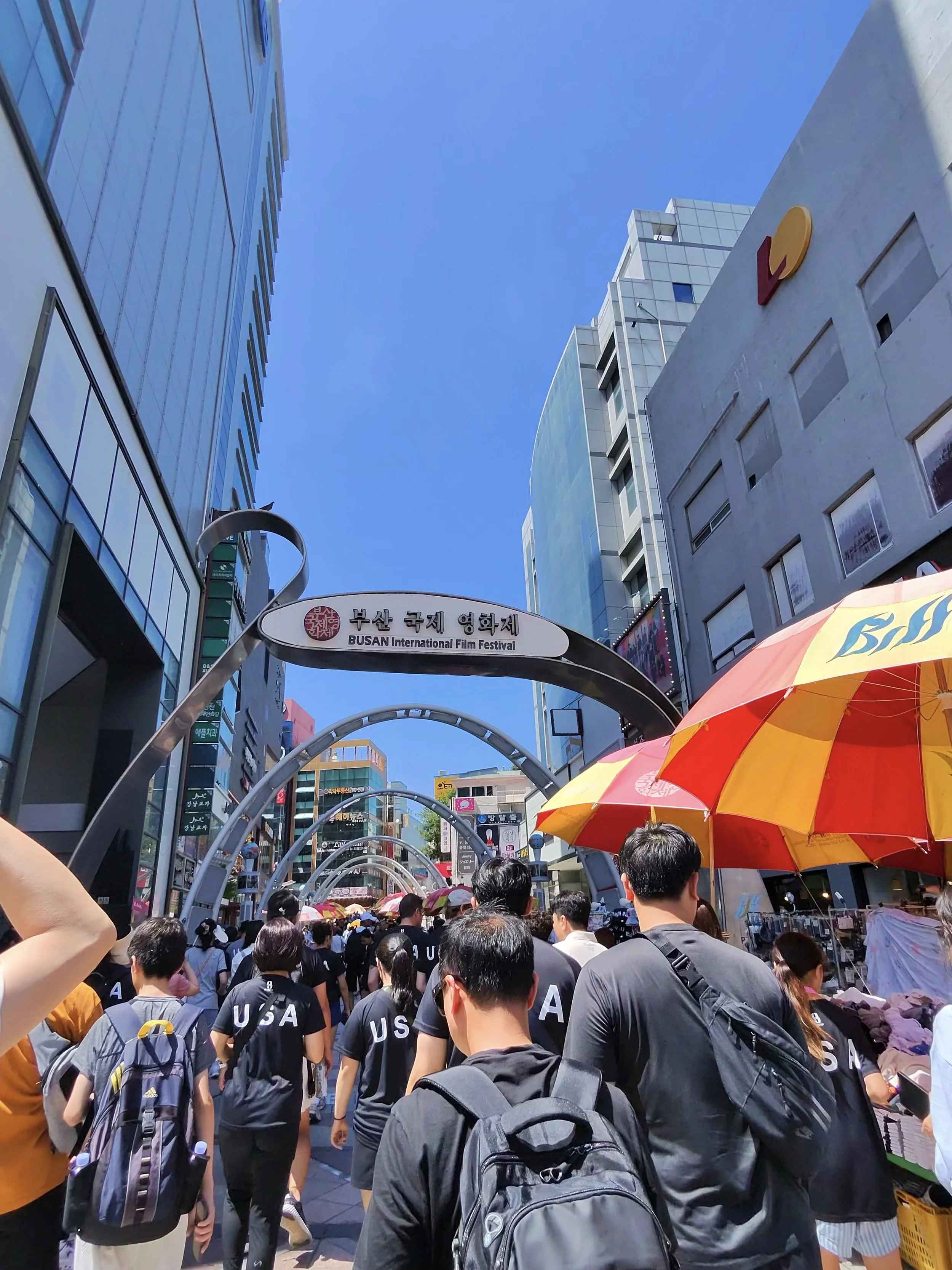 Crowd of people walking under umbrellas on a sunny day in an urban area, with tall buildings and signs in Korean and English, including a sign for the Busan International Film Festival.