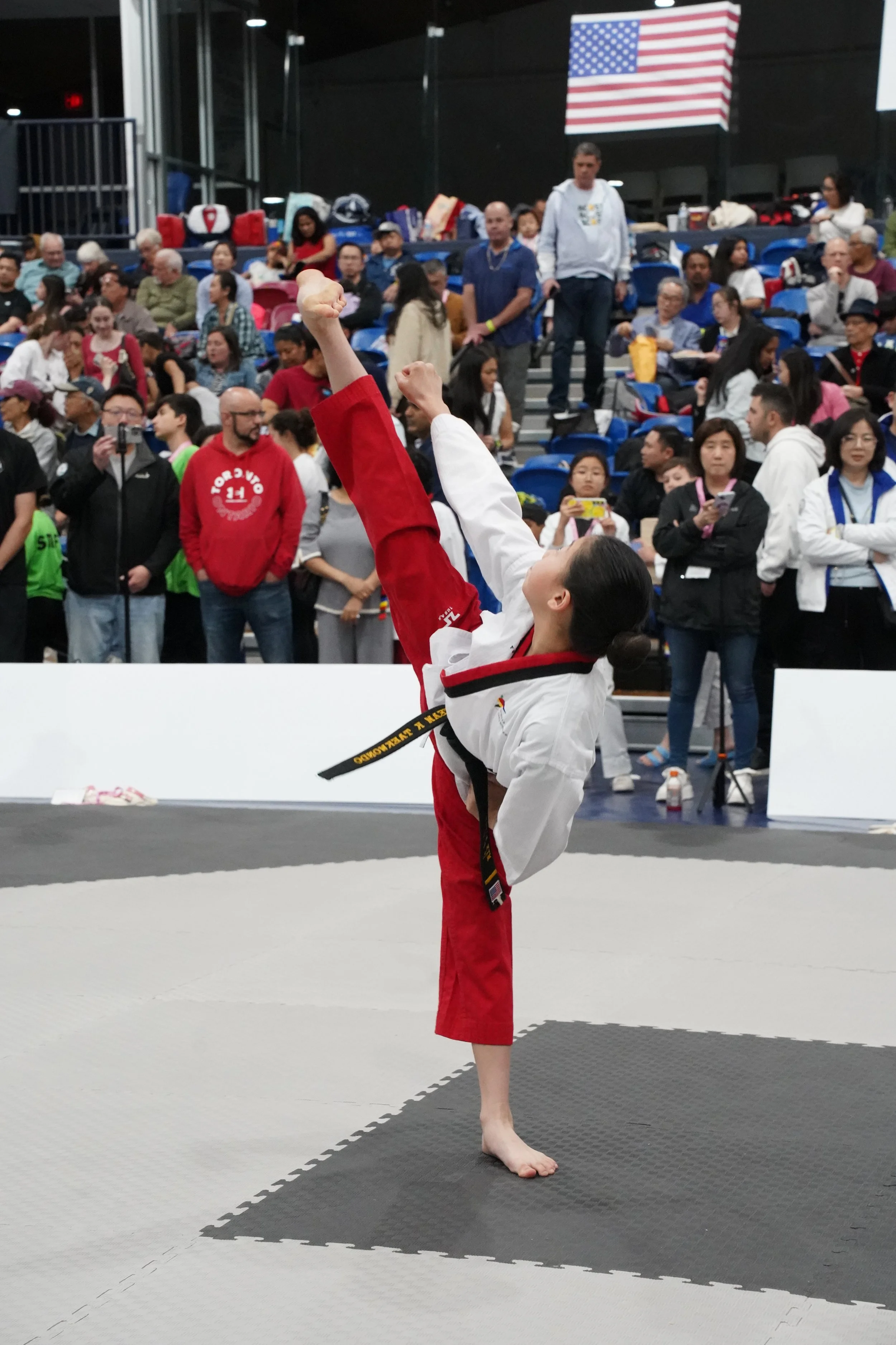 A young girl in a red and white martial arts uniform standing barefoot on a mat in a gymnasium, with her arm raised in a fist. There are many people in the background, including spectators and other participants, with an American flag displayed on the wall.