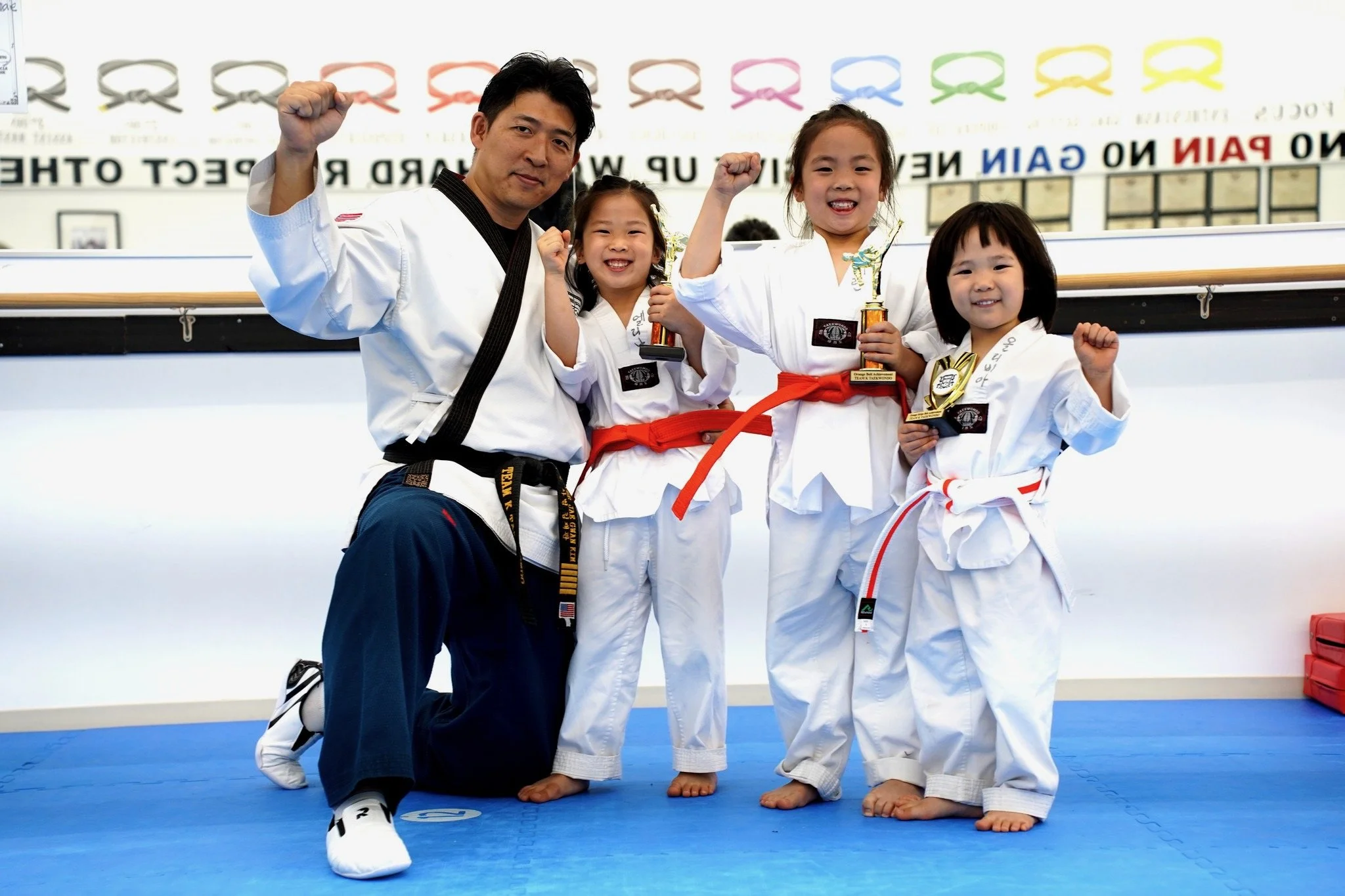 A martial arts instructor and three young girls, all wearing white uniforms and colorful belts, celebrating with fists raised in a dojo. The girls are smiling and holding trophies, indicating a competition or achievement.