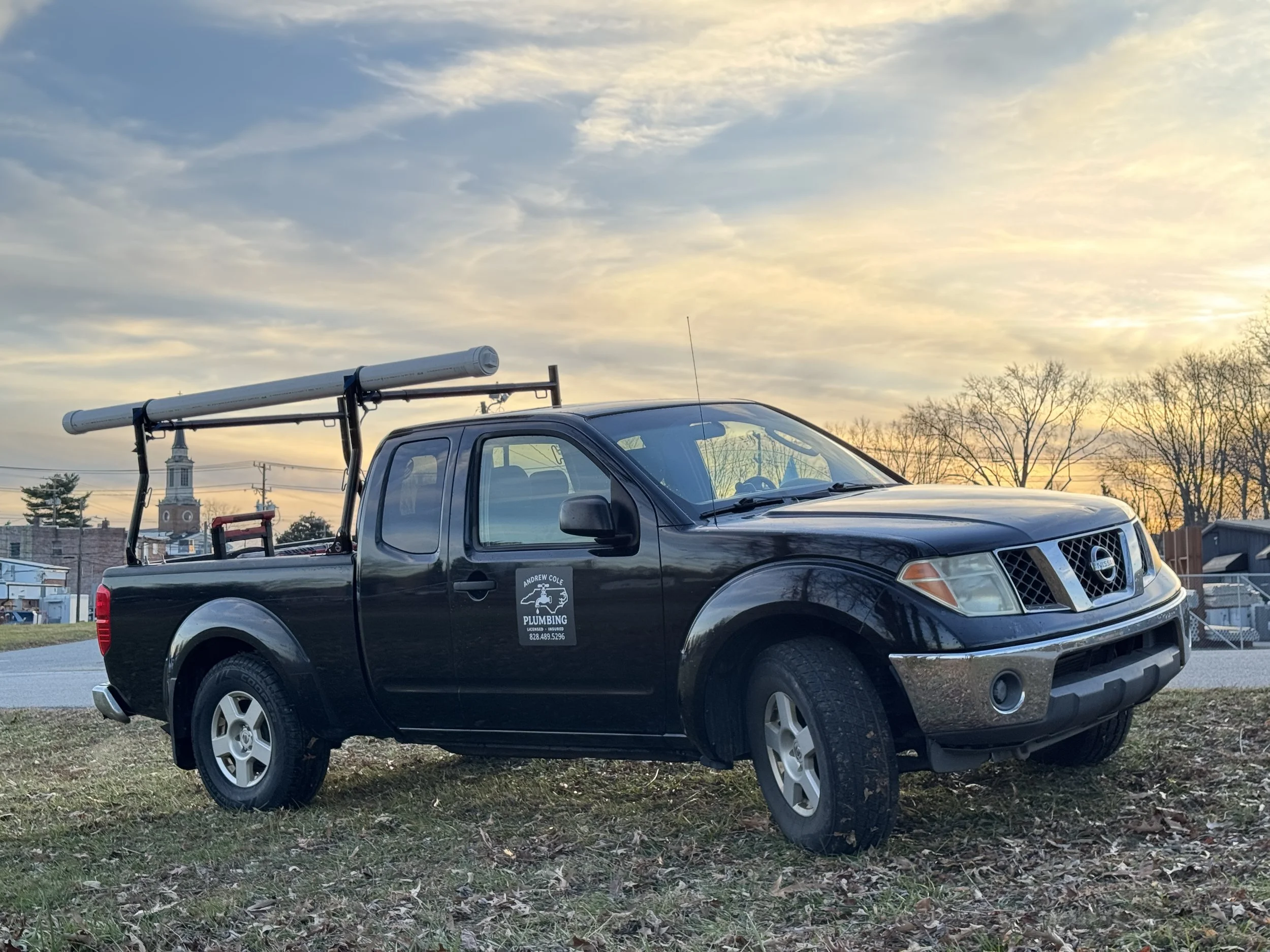 Black pickup truck with plumbing company logo parked on grassy area at sunset, with trees and buildings in the background.