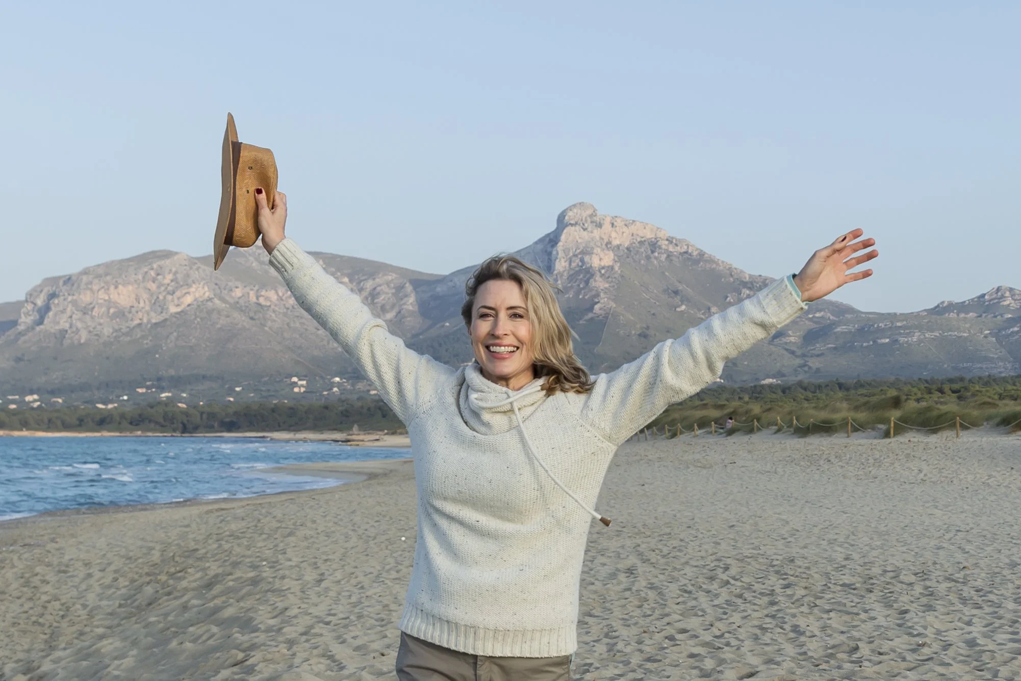 A woman standing on the beach with her arms raised, holding a hat in her right hand, smiling, with mountains and the ocean in the background during daytime.