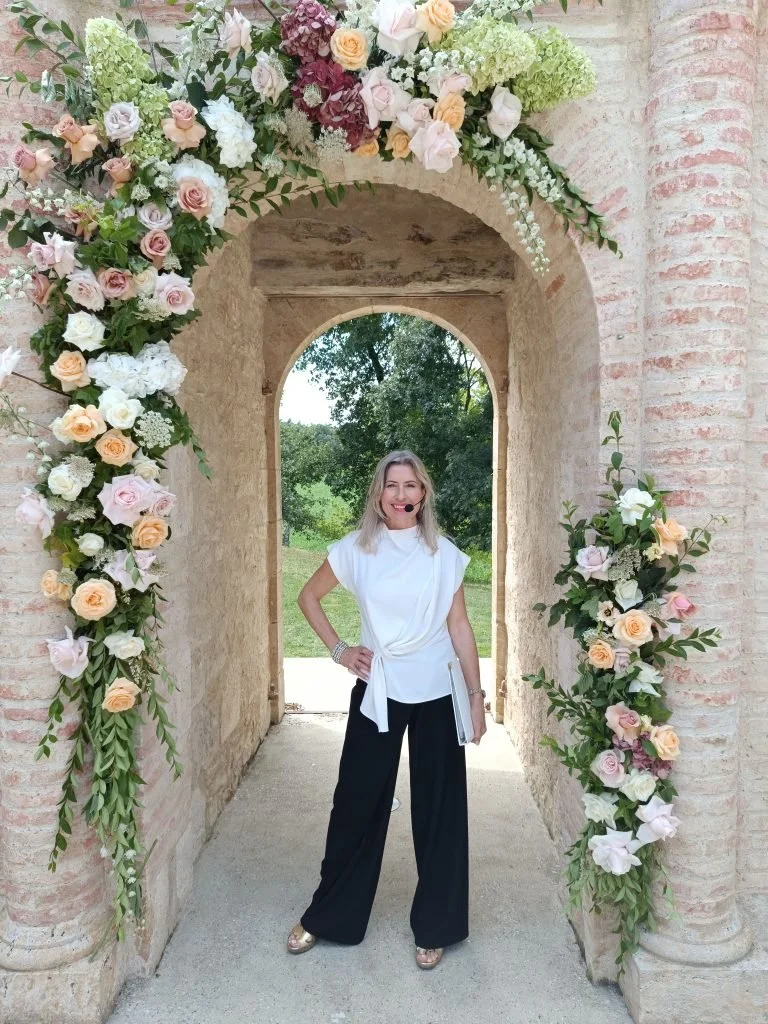 Laura The Chateau Celebrant standing under an arch at Chateau Englain decorated with flowers, outdoors on a sunny day, smiling.