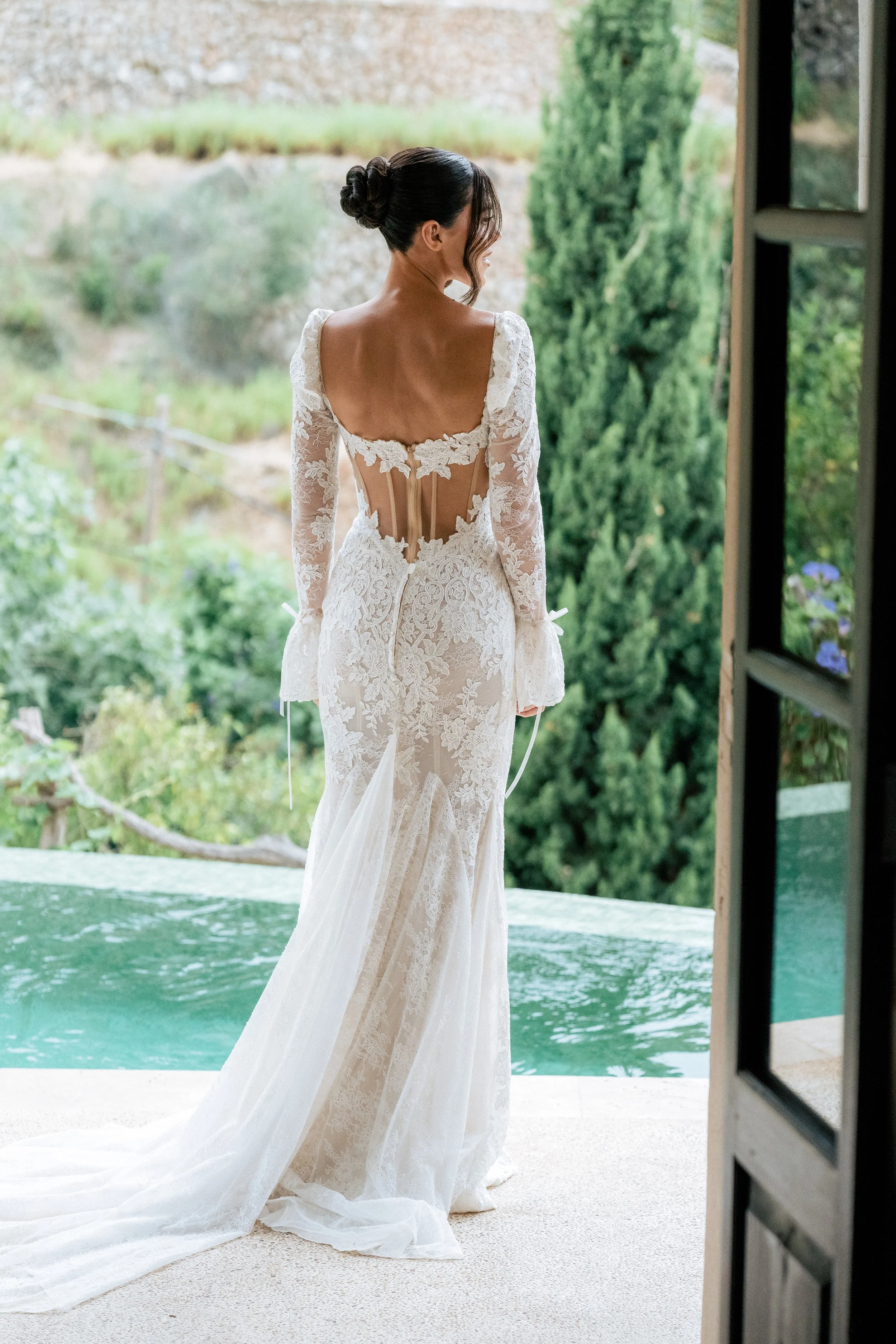 Woman in a white lace wedding dress standing at an open door, looking outside at a lush green landscape with trees and a pond.