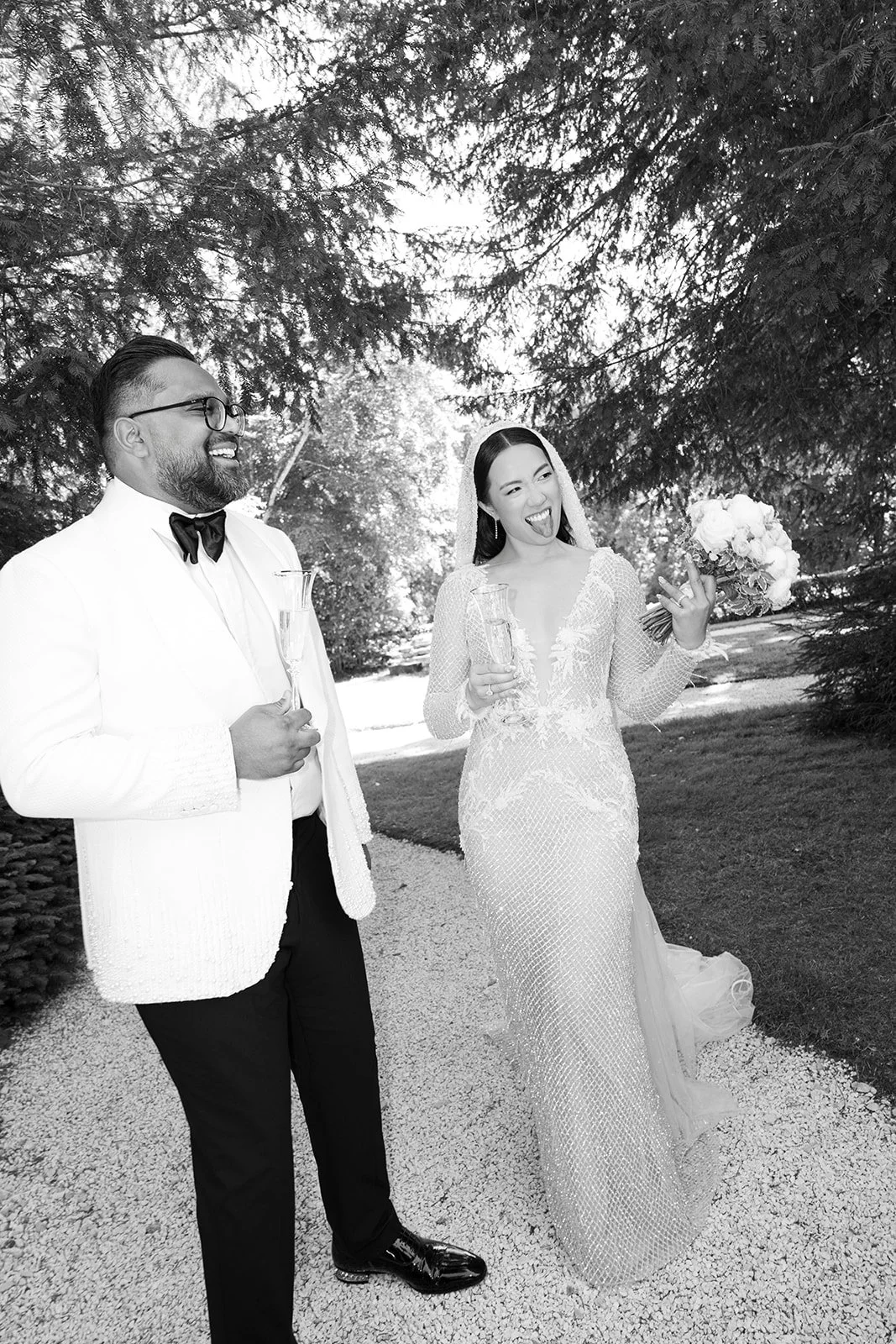A bride and groom at their wedding celebration outdoors, smiling and holding glasses, with trees in the background.