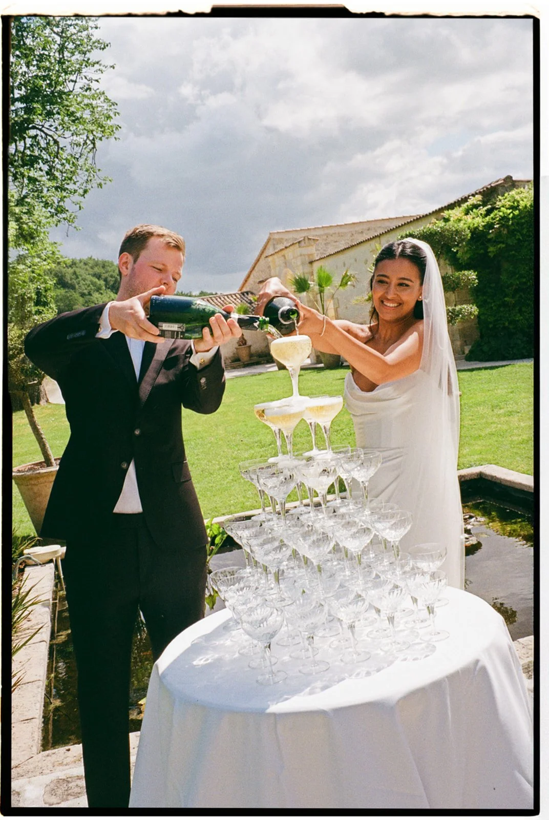 A newlywed couple celebrating during their wedding reception outdoors, pouring champagne into a pyramid of glasses, with greenery and a building in the background.