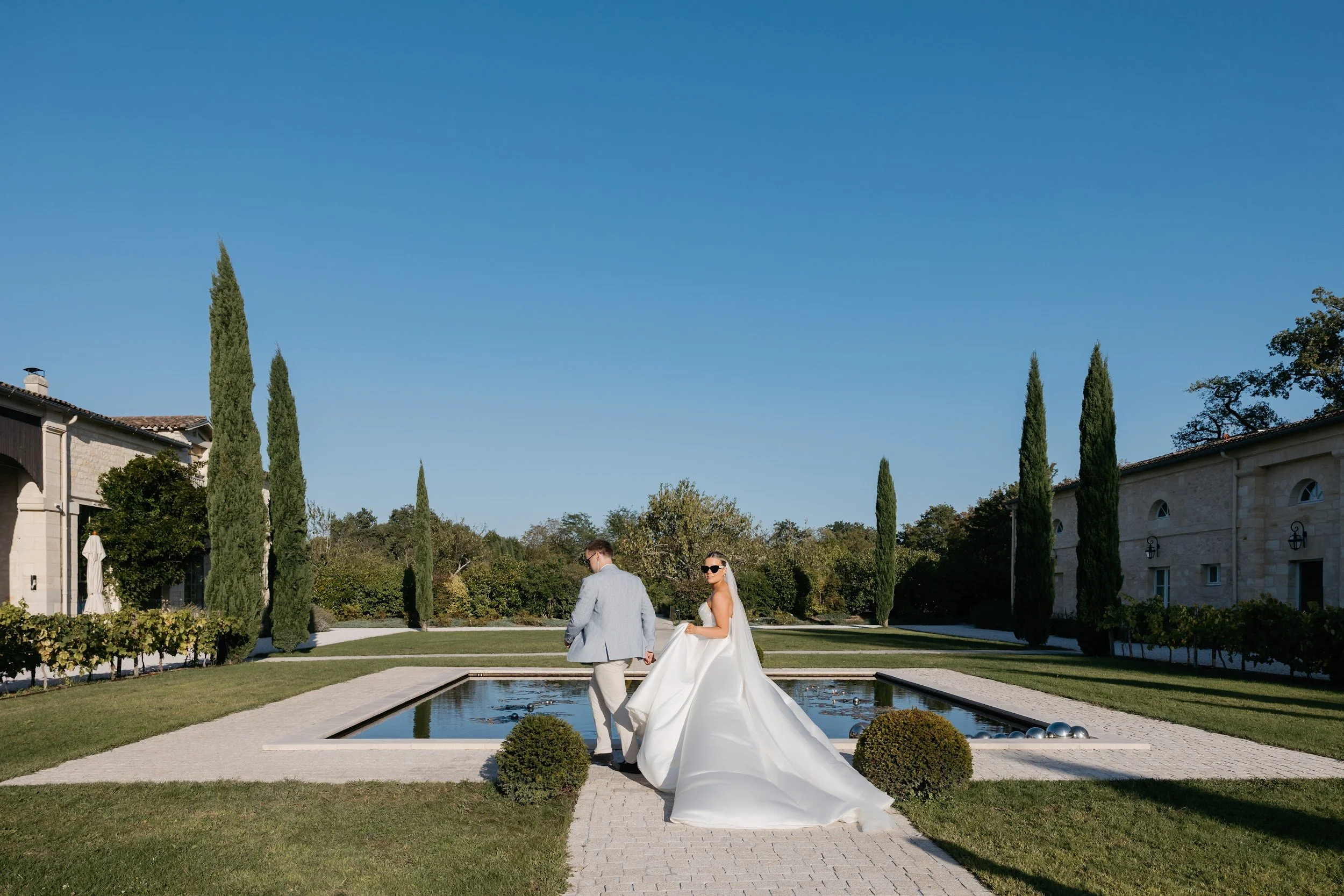 A newlywed couple holding hands and walking near a rectangular pond on a sunny day, surrounded by tall cypress trees and elegant buildings, with the bride wearing a white wedding gown and sunglasses.