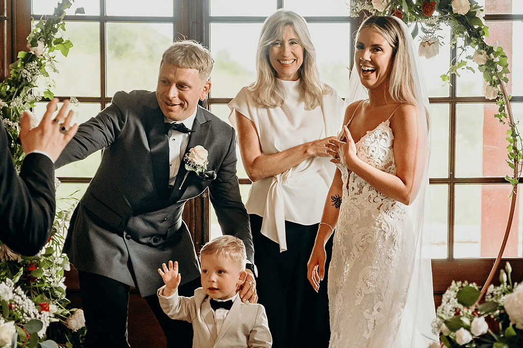 A bride, groom, and family members celebrate at a wedding. The bride is in a white lace wedding dress, the groom in a grey tuxedo, a woman in a white top, and a young boy in a tuxedo, with a floral decorated backdrop.