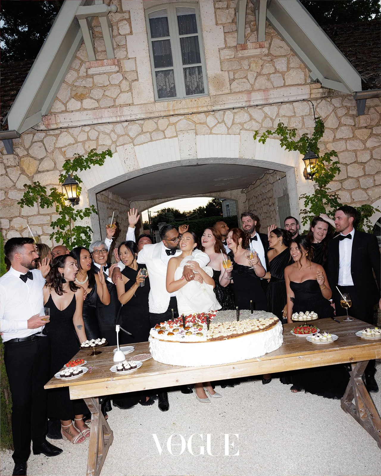 A large group of people dressed in formal attire celebrating at a wedding reception outdoors, with a bride and groom at the center near a large cake on a wooden table.