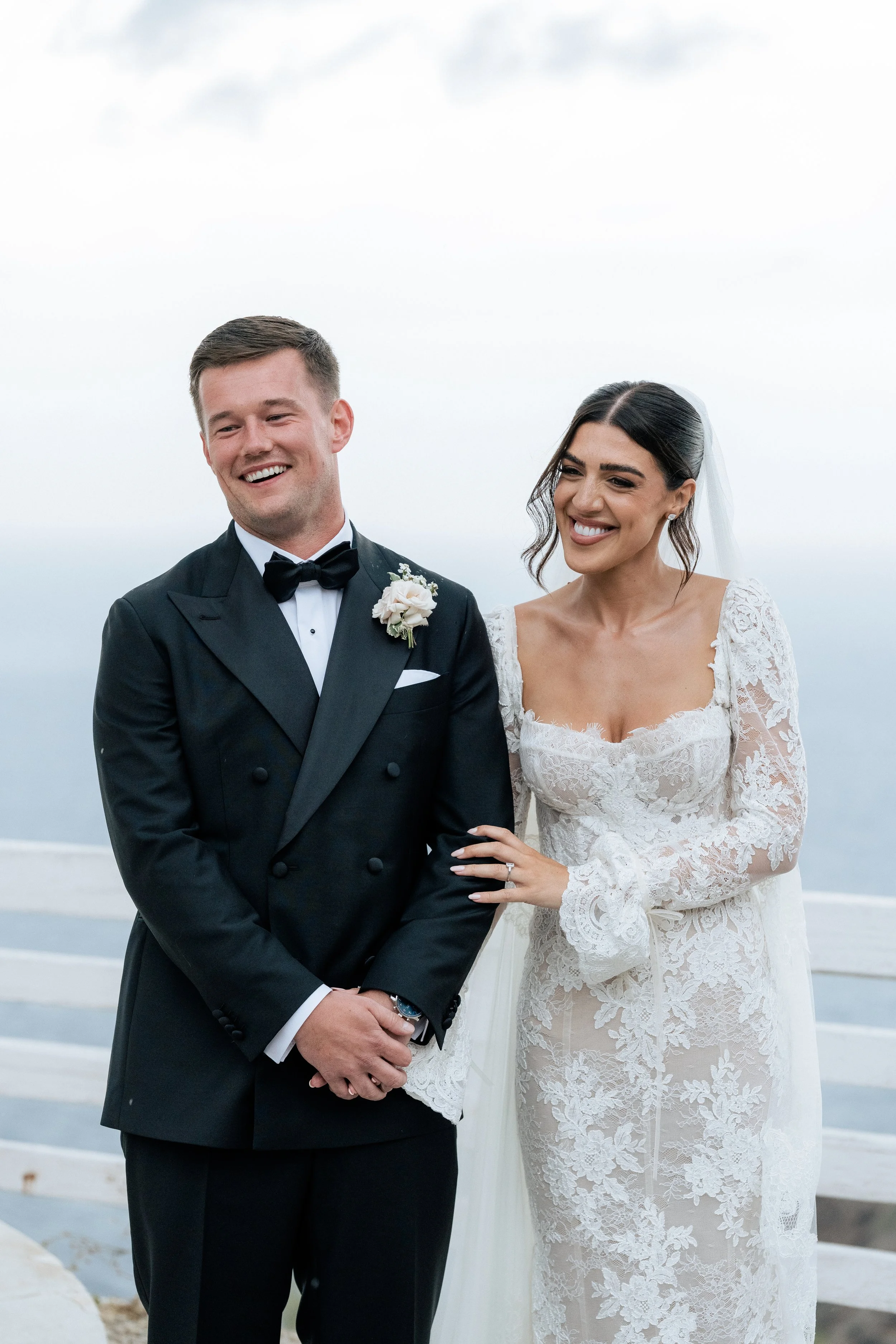 A bride and groom smiling at their wedding ceremony outdoors, with the ocean in the background. The groom is wearing a black tuxedo with a bow tie and boutonniere, and the bride is in a white lace wedding gown with a veil.