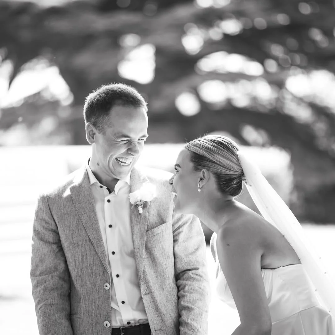 A bride and groom laughing and smiling together outdoors on their wedding day, with trees and natural light in the background.