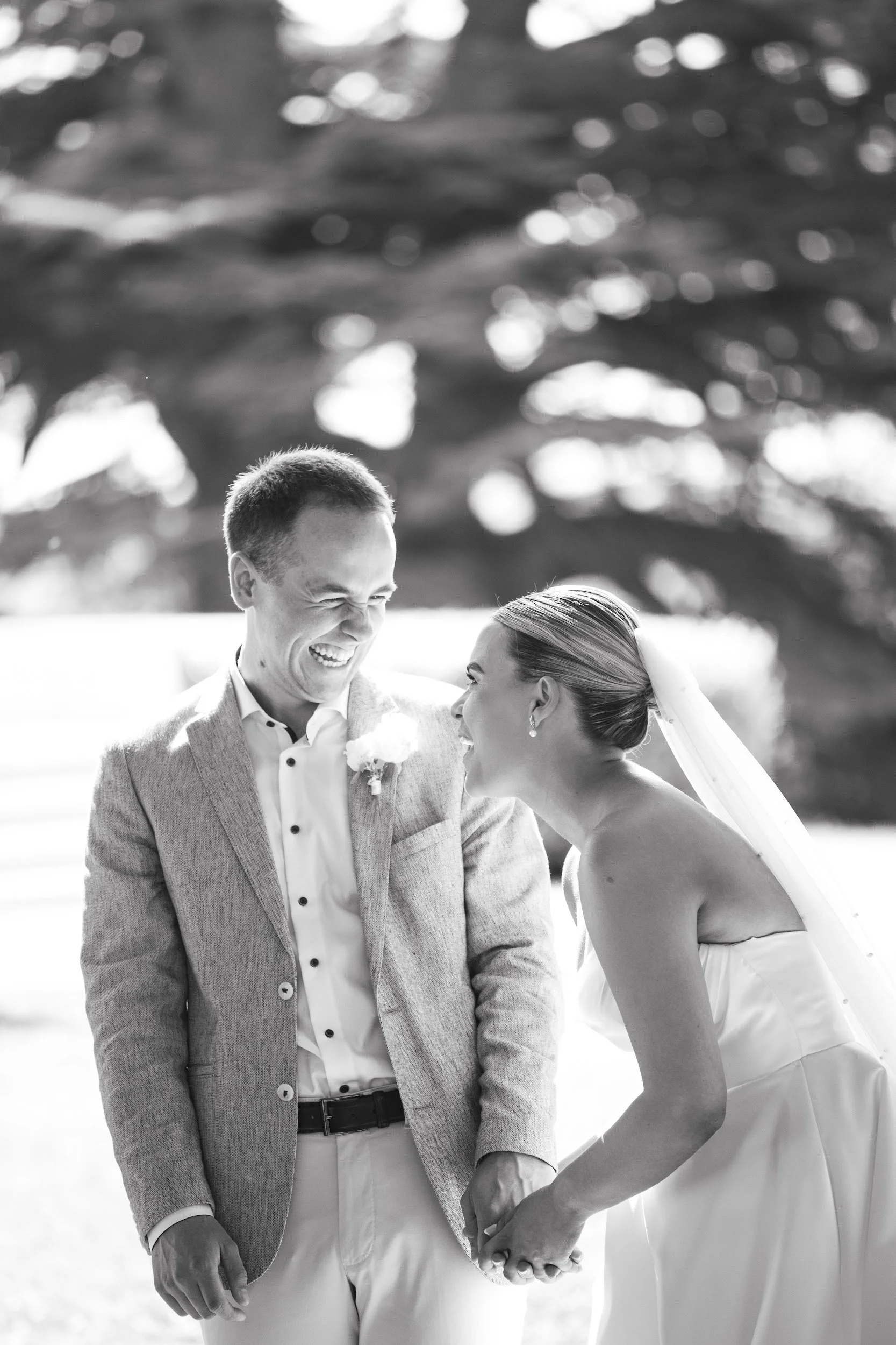 Black and white photo of a bride and groom holding hands and smiling at each other outdoors.