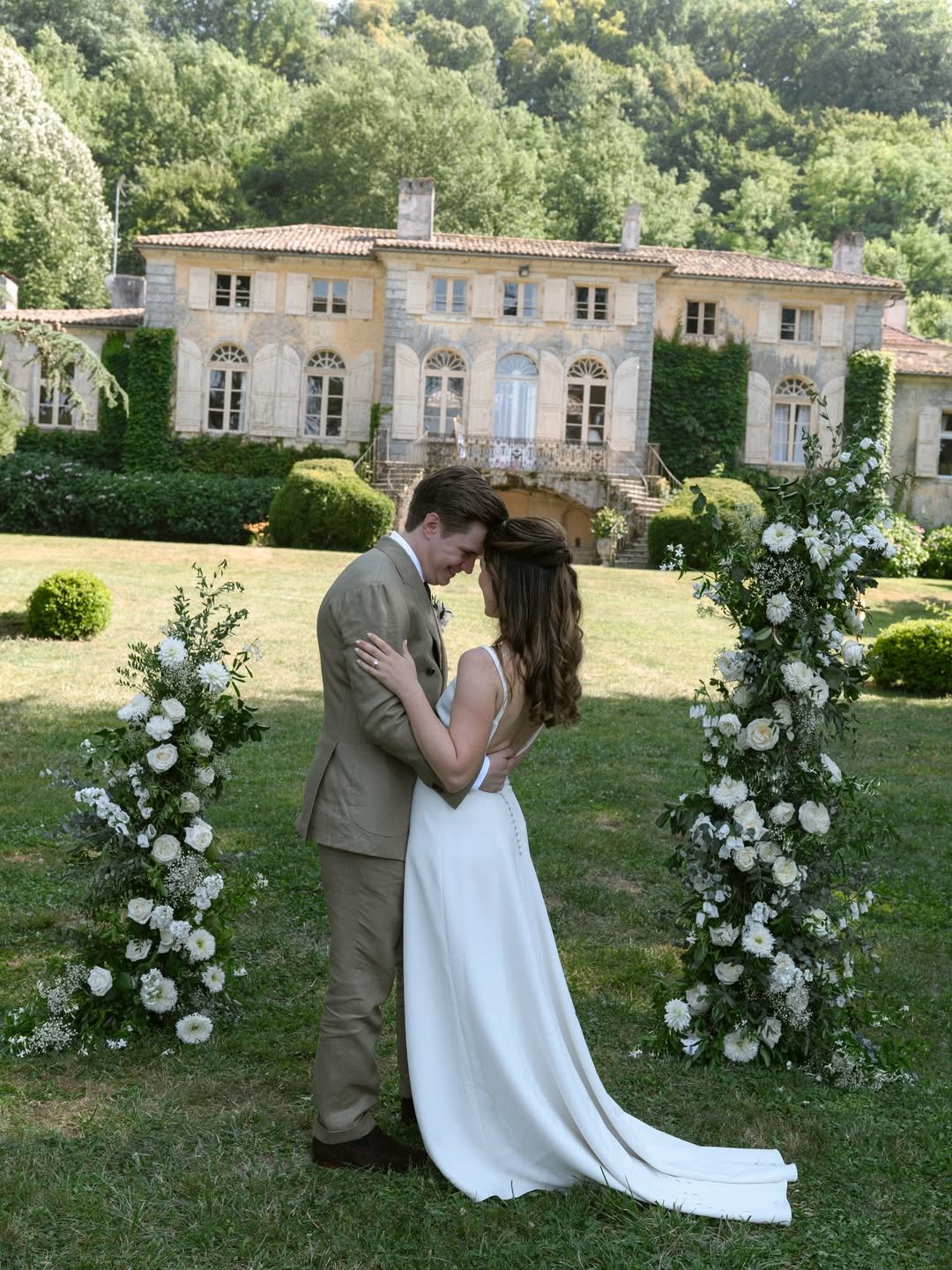 A bride and groom embrace at an outdoor wedding ceremony, standing between two floral arrangements with a large estate or mansion in the background.
