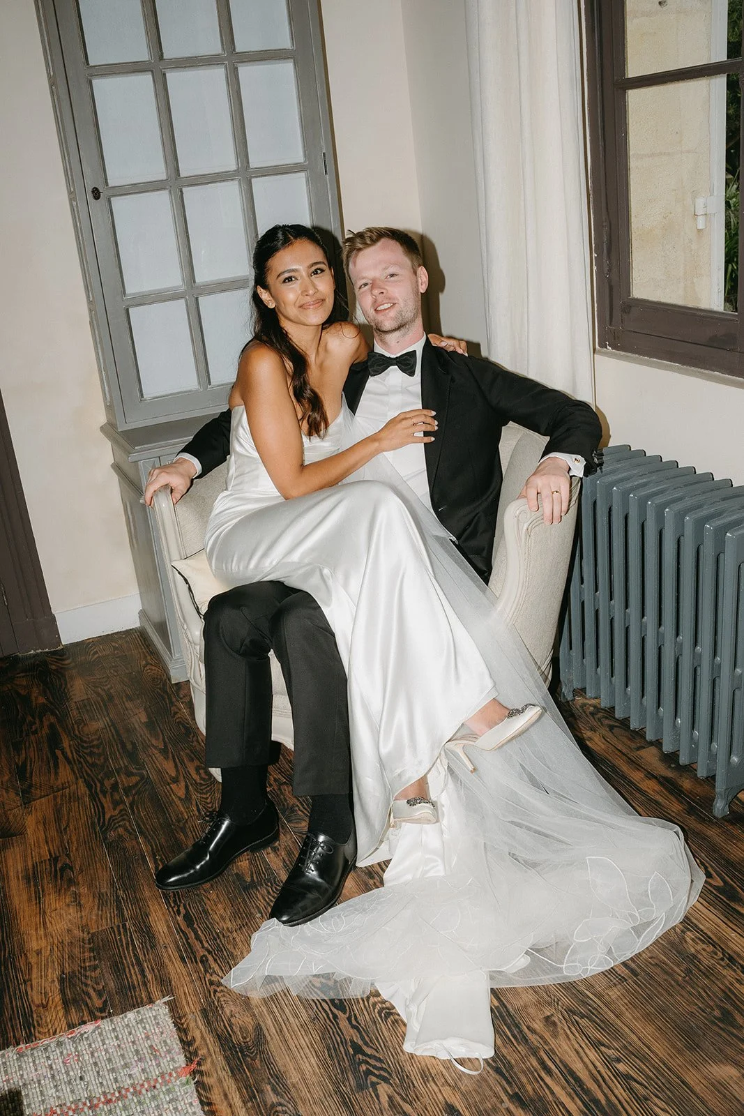 A bride and groom sitting together on a sofa. The bride is in a white wedding gown, and the groom is in a black tuxedo. They are smiling and posing for the camera in a room with a wooden floor, a radiator, and a window.