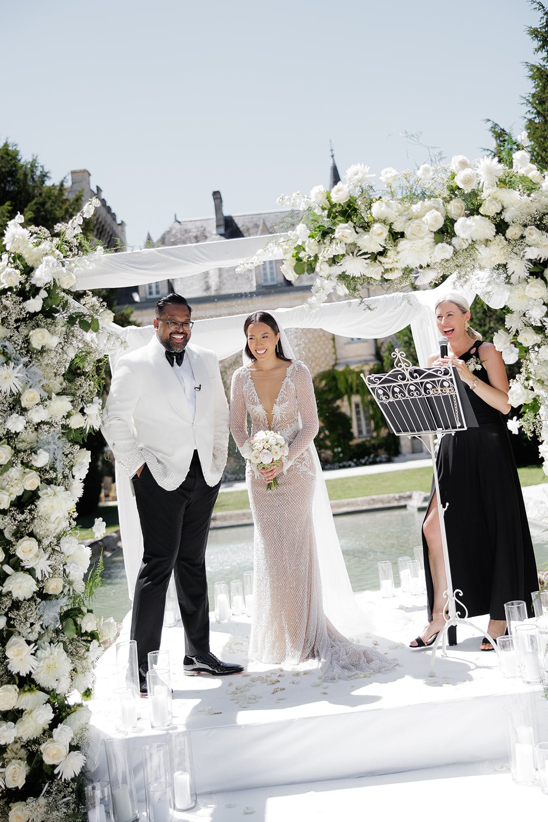 A wedding ceremony taking place outdoors under a floral arch, with a bride and groom standing together, and Laura the Chateau Celebrant officiating.
