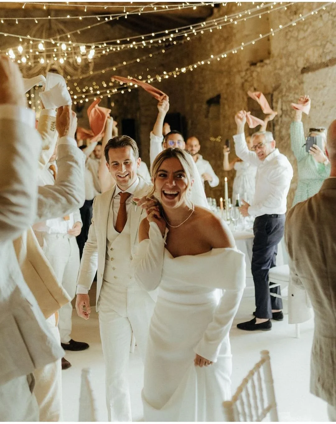 People celebrating at a wedding reception with string lights and joyful expressions.