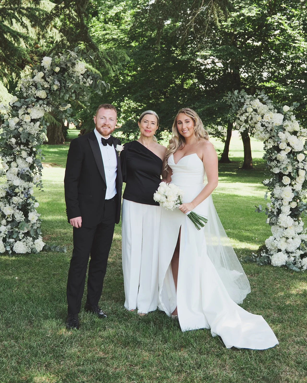 A bride and groom standing with  under a floral arch at an outdoor wedding. The groom is in a black tuxedo, the bride is in a white wedding dress holding a bouquet, and the woman is in a black and white outfit. Green trees and grass form the background.