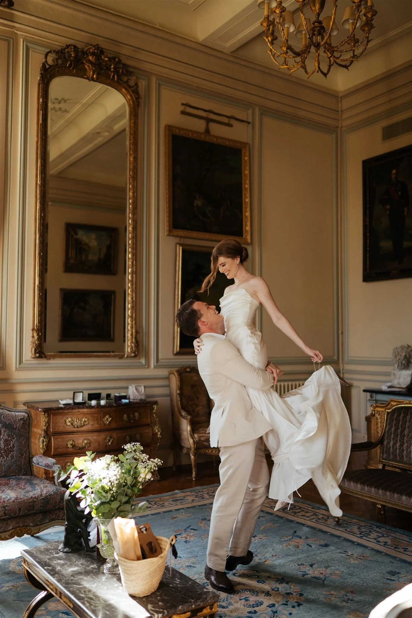 A man in a white suit lifting a woman in a white wedding dress in a fancy, ornate room with paintings, a chandelier, and vintage furniture.