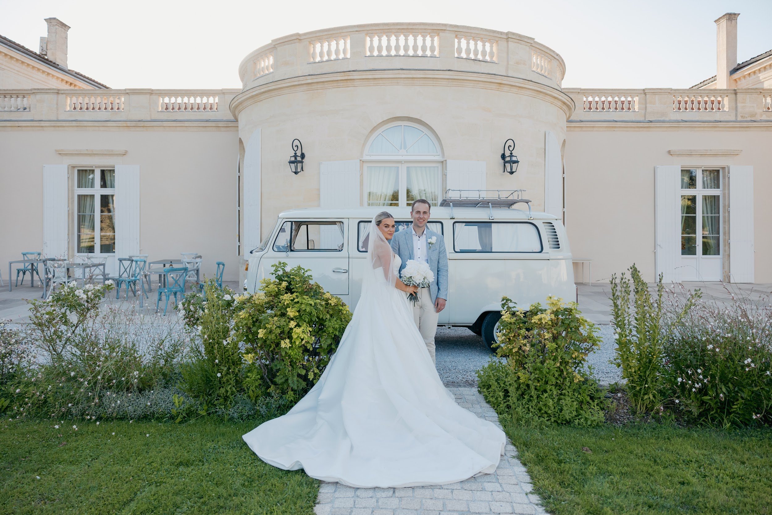 Bride and groom standing in front of a vintage van with a large house behind them, surrounded by greenery and outdoor seating.
