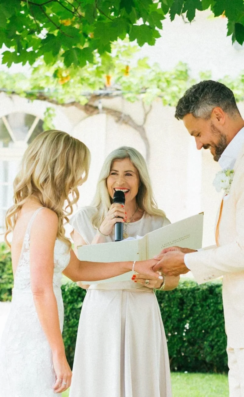 Laura The Chateau Celebrant officiating a wedding ceremony outdoors under green leafy trees, a bride and groom exchanging vows. At Chateau Soulac.