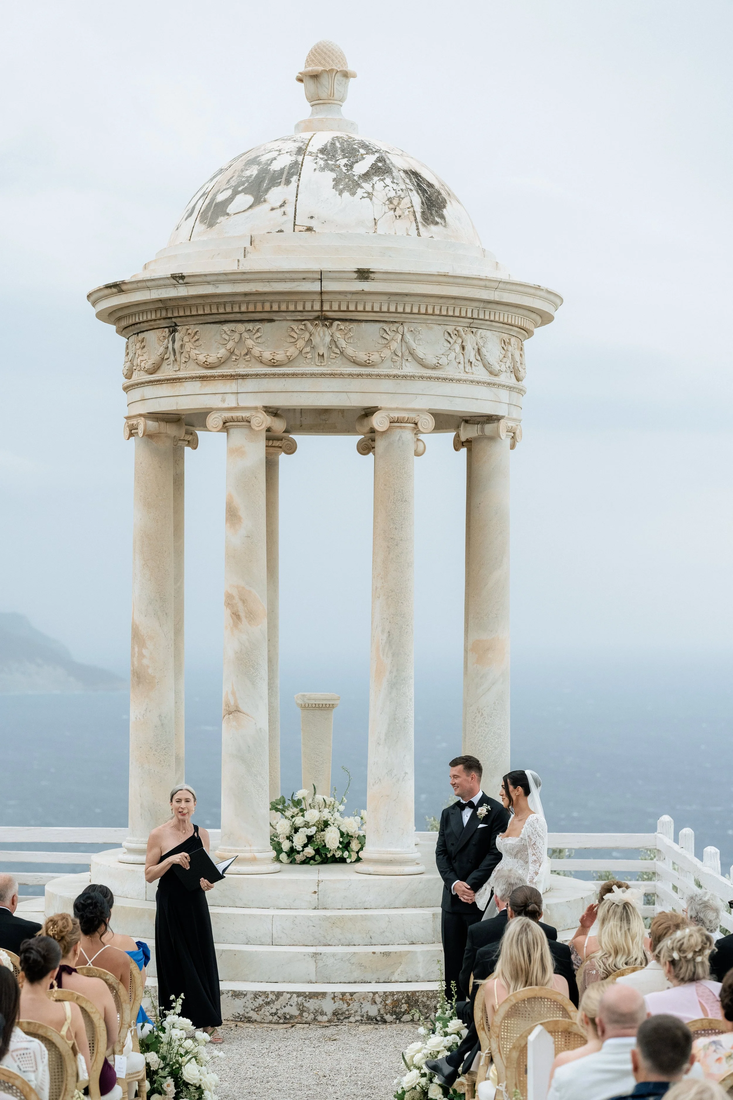 A wedding ceremony taking place at Son Marroig in Malloca outdoors under a marble gazebo with columns, overlooking the ocean and coastline, with guests seated and Laura Shuckburgh officiating.
