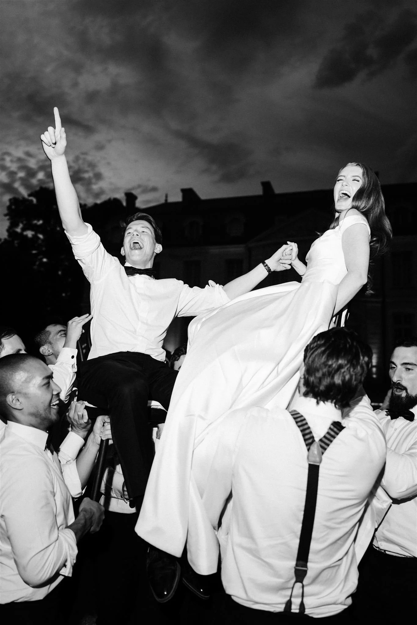 A group of people celebrating outdoors at night, lifting a woman in a white dress and a man in a white shirt and bow tie on a chair, all smiling and laughing.