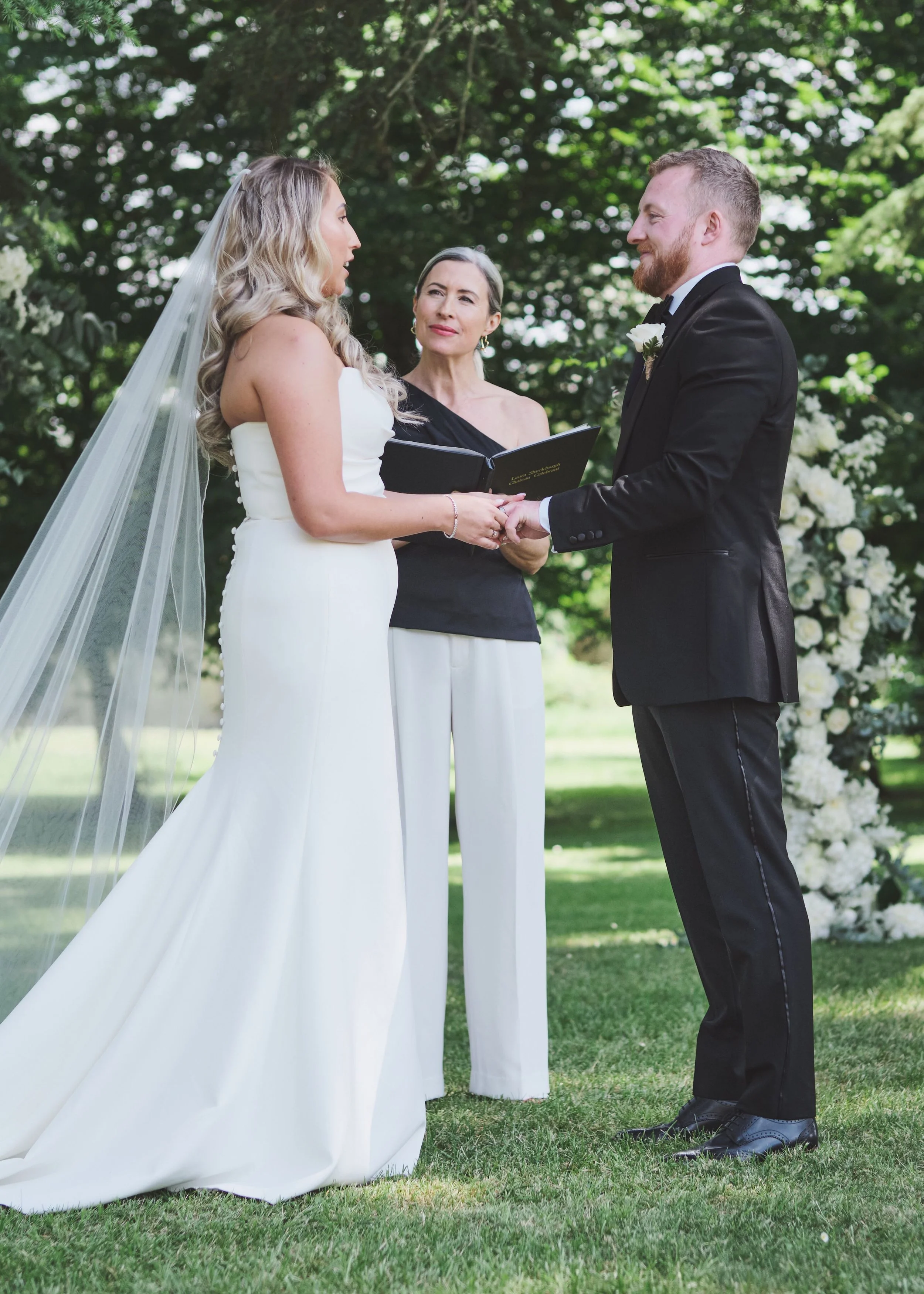 A bride and groom holding hands during their outdoor wedding ceremony, facing each other, with an officiant standing behind them, in a lush green setting with a floral arrangement.